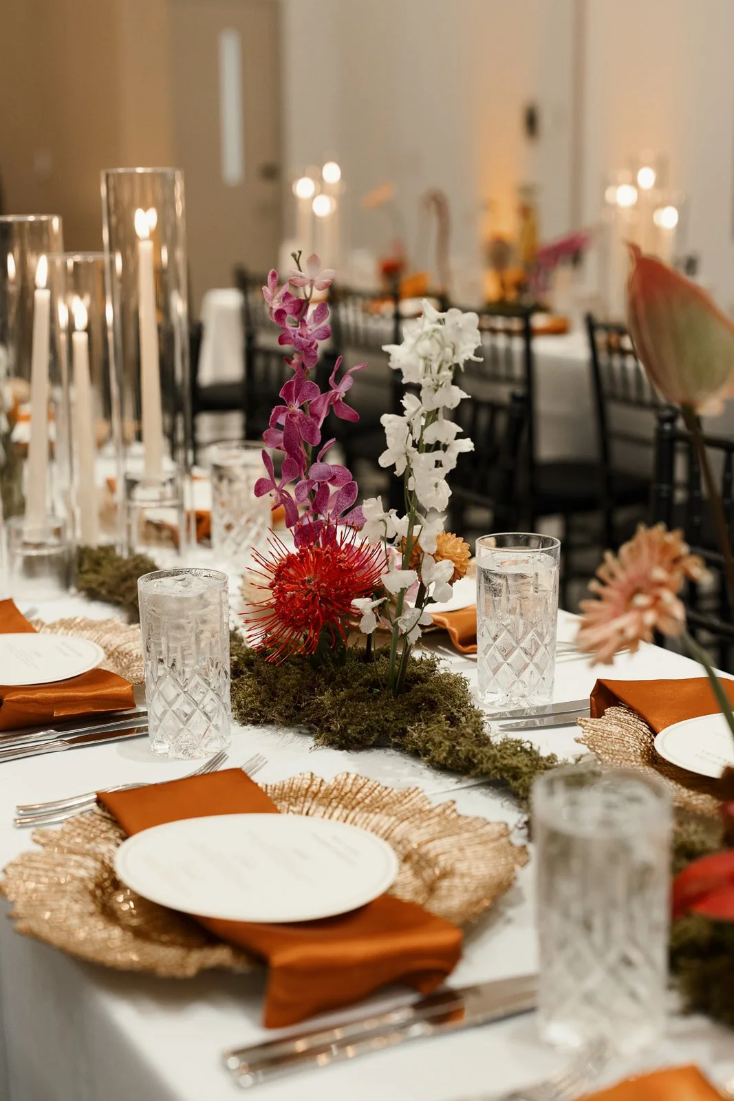 Elegant banquet table setting with floral arrangement, candles, and placed plates with orange napkins.