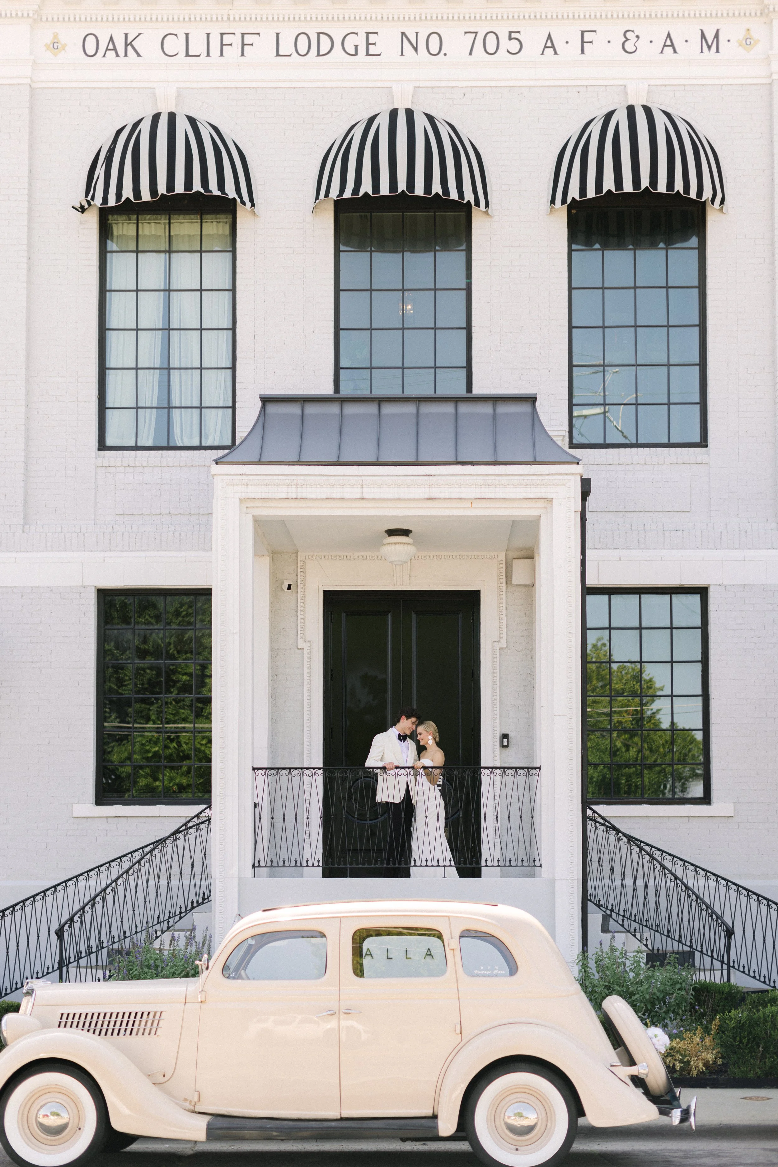 A couple dressed in vintage wedding attire standing on the front porch of a white brick building with black doors and large windows, with three black-and-white striped awnings and a vintage beige car parked in front.