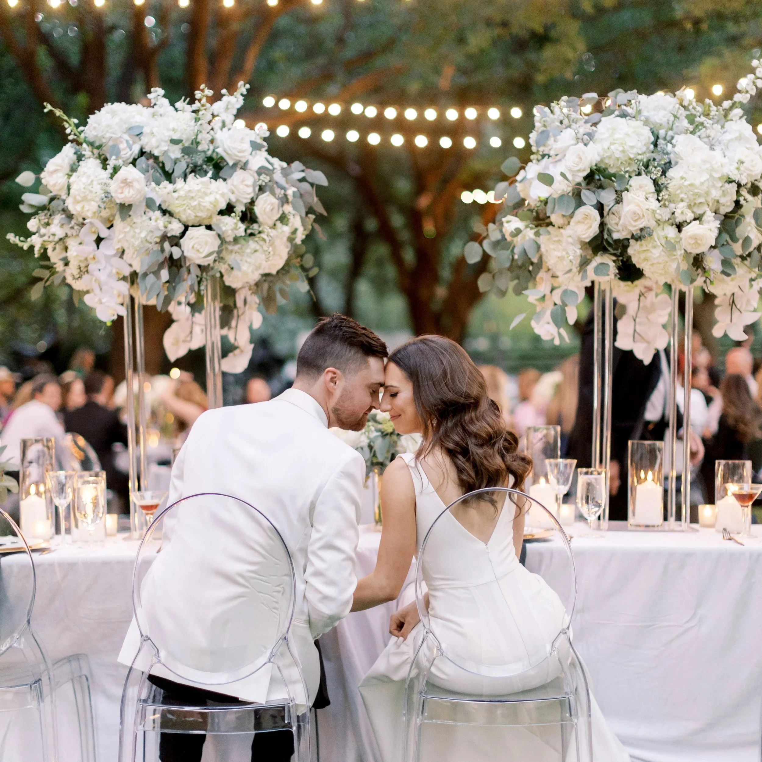 A bride and groom sit close together with foreheads touching at a wedding reception table outdoors, surrounded by tall floral arrangements, candles, and guests in the background.