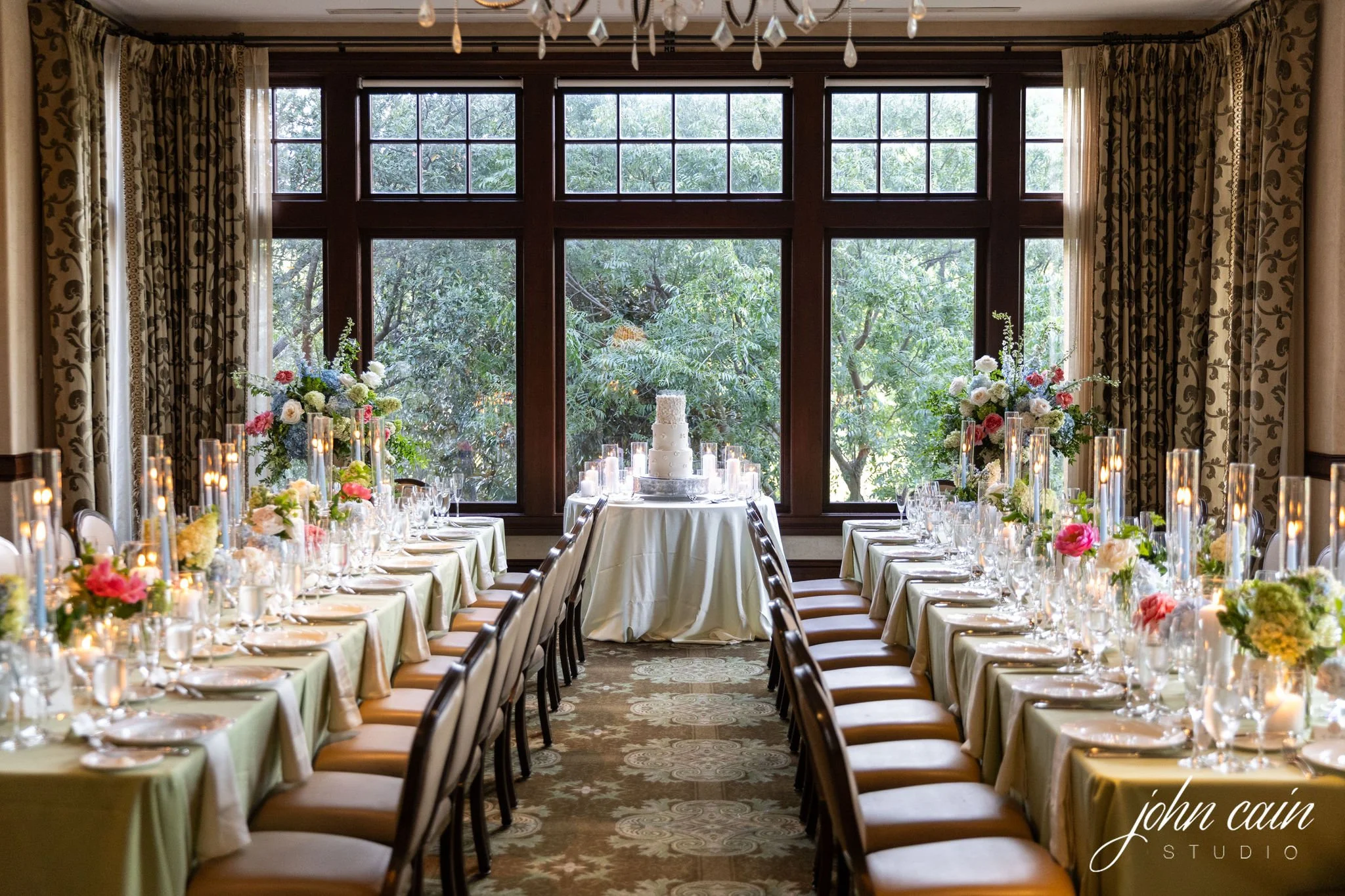 Elegant wedding reception setup with long tables covered in cream tablecloths, decorated with floral centerpieces and tall glass candle holders, and a wedding cake on a small table at the back near a large window with trees outside.