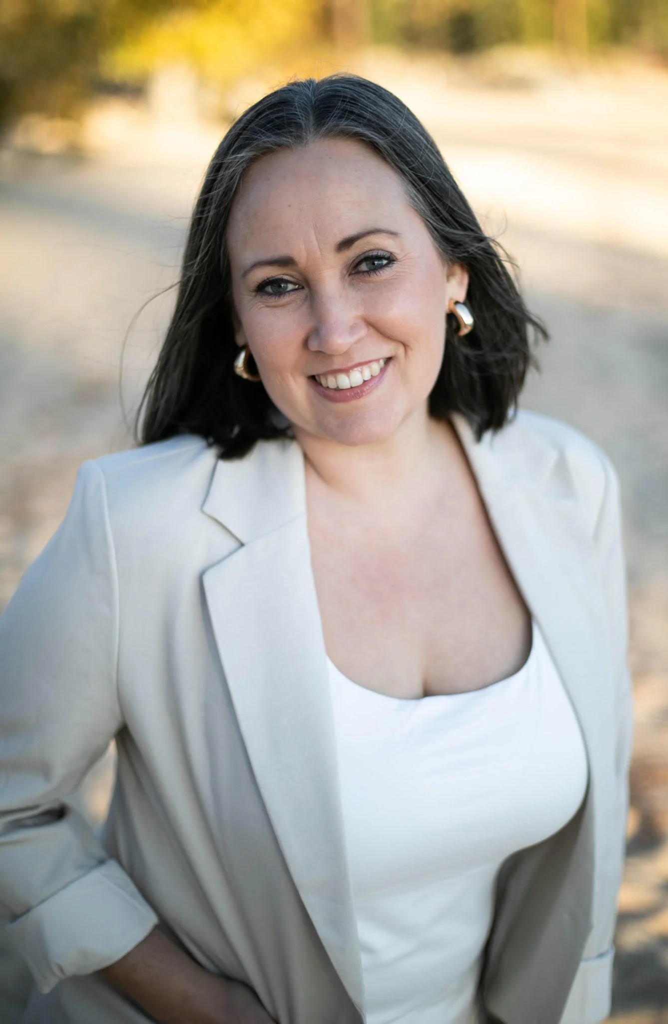 A woman with shoulder-length dark hair, blue eyes, and earrings, smiling outdoors during daylight, wearing a white top and light-colored blazer.