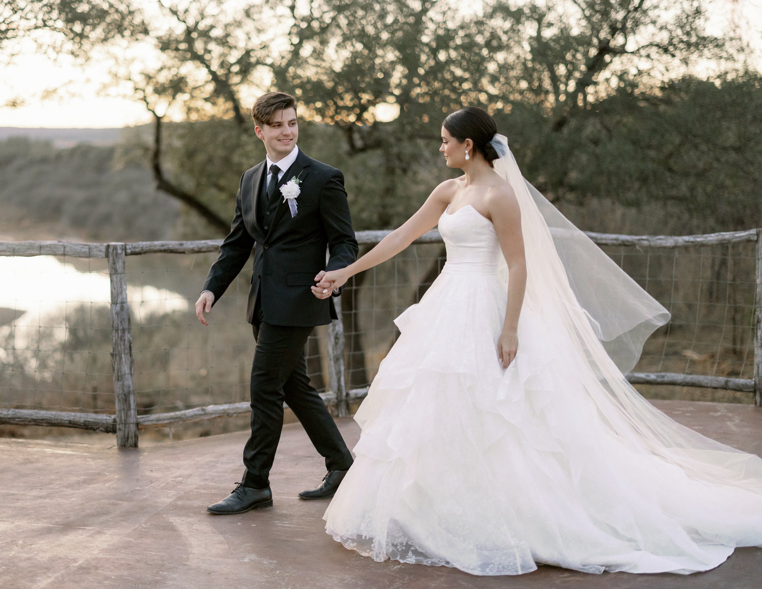 A bride and groom holding hands and walking outdoors during their wedding, with trees and a rustic wooden fence in the background at sunset.