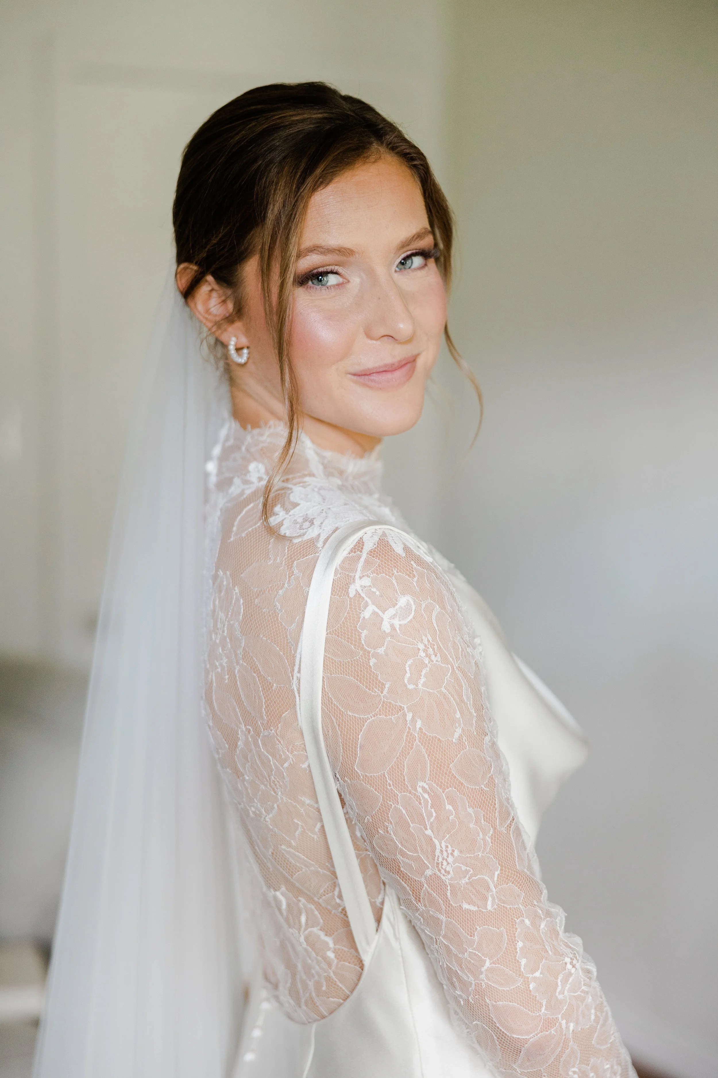 A woman in a wedding dress with lace sleeves, smiling softly, with makeup and loose hair, in an indoor setting.