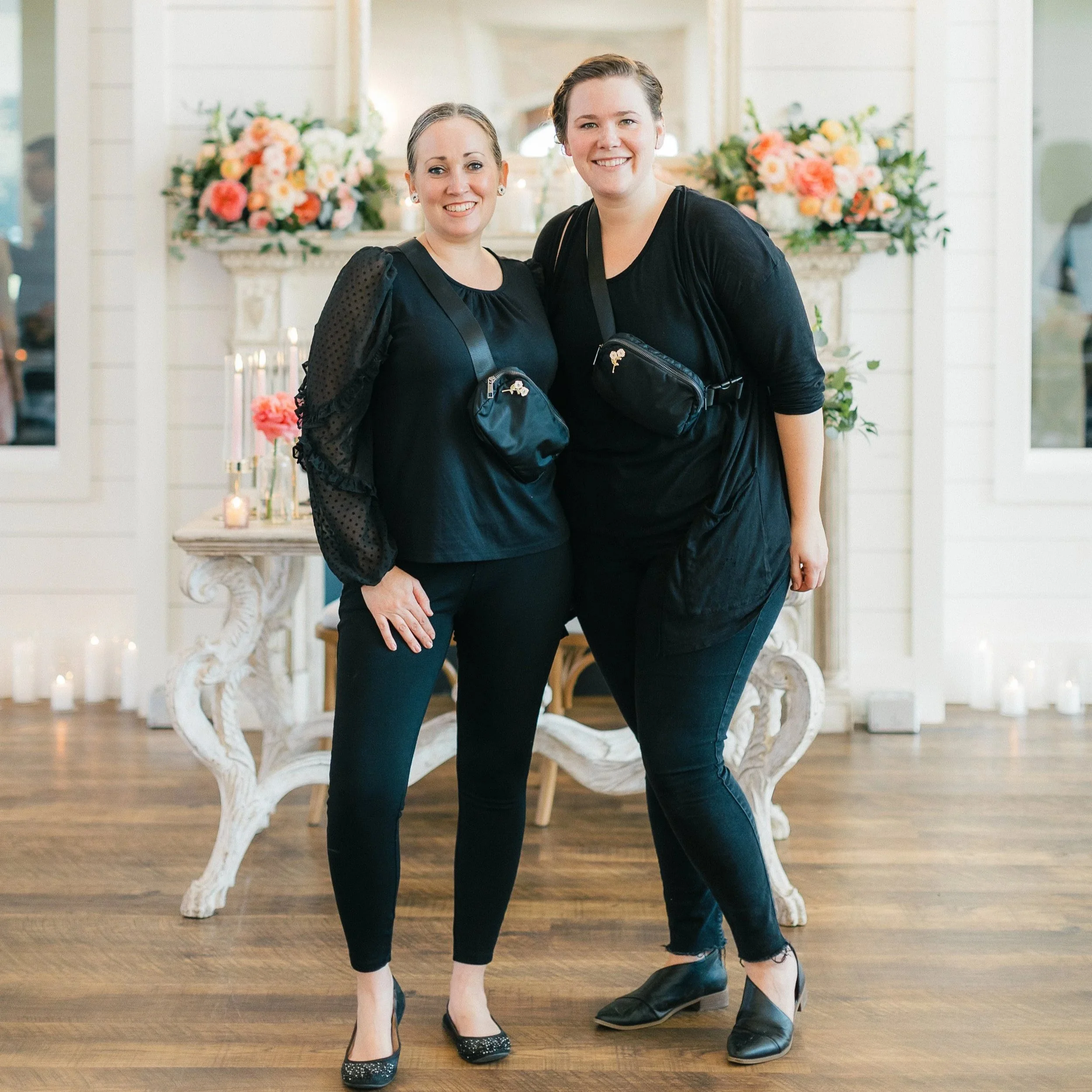 Two women dressed in black standing inside a decorated room with flowers and candles, smiling at the camera.