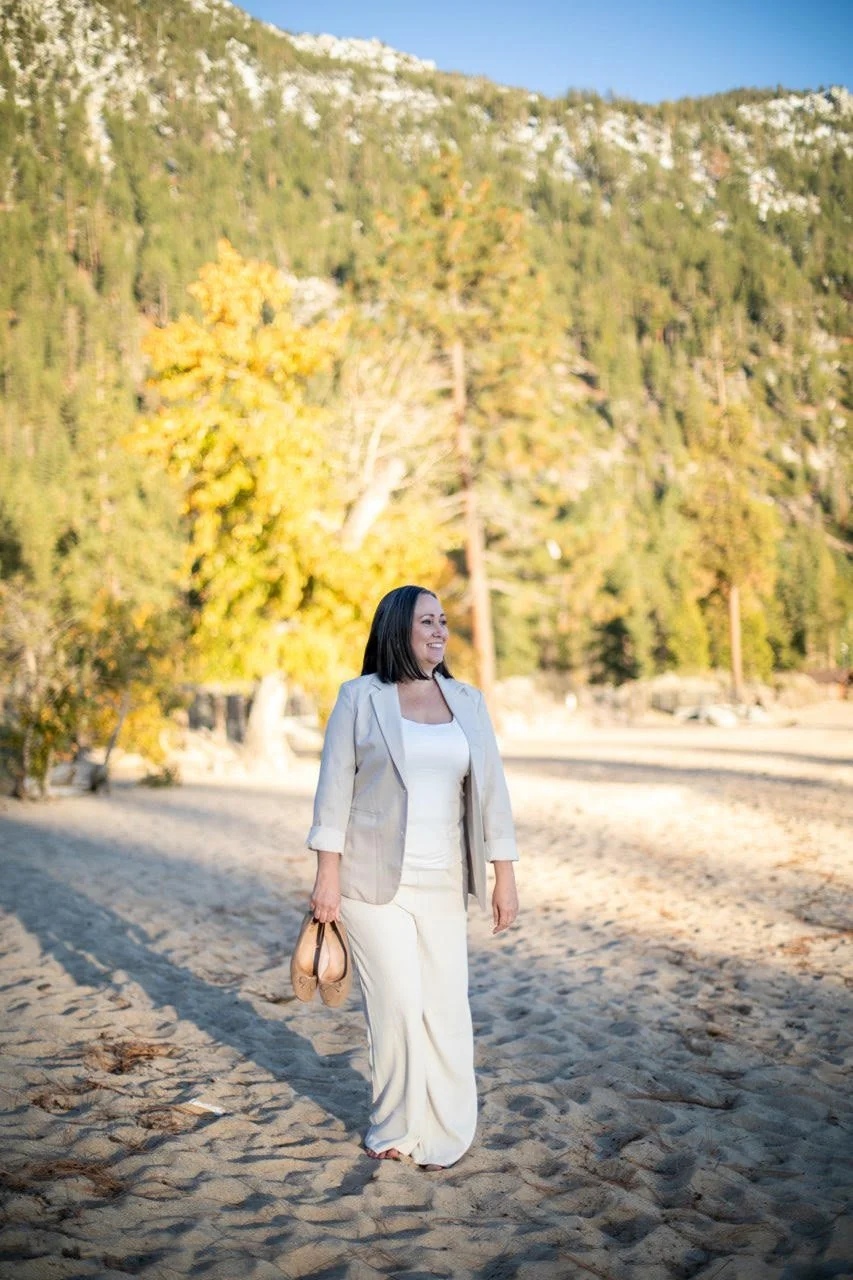 Woman in a light-colored suit walking on sandy beach with trees and mountains in the background.