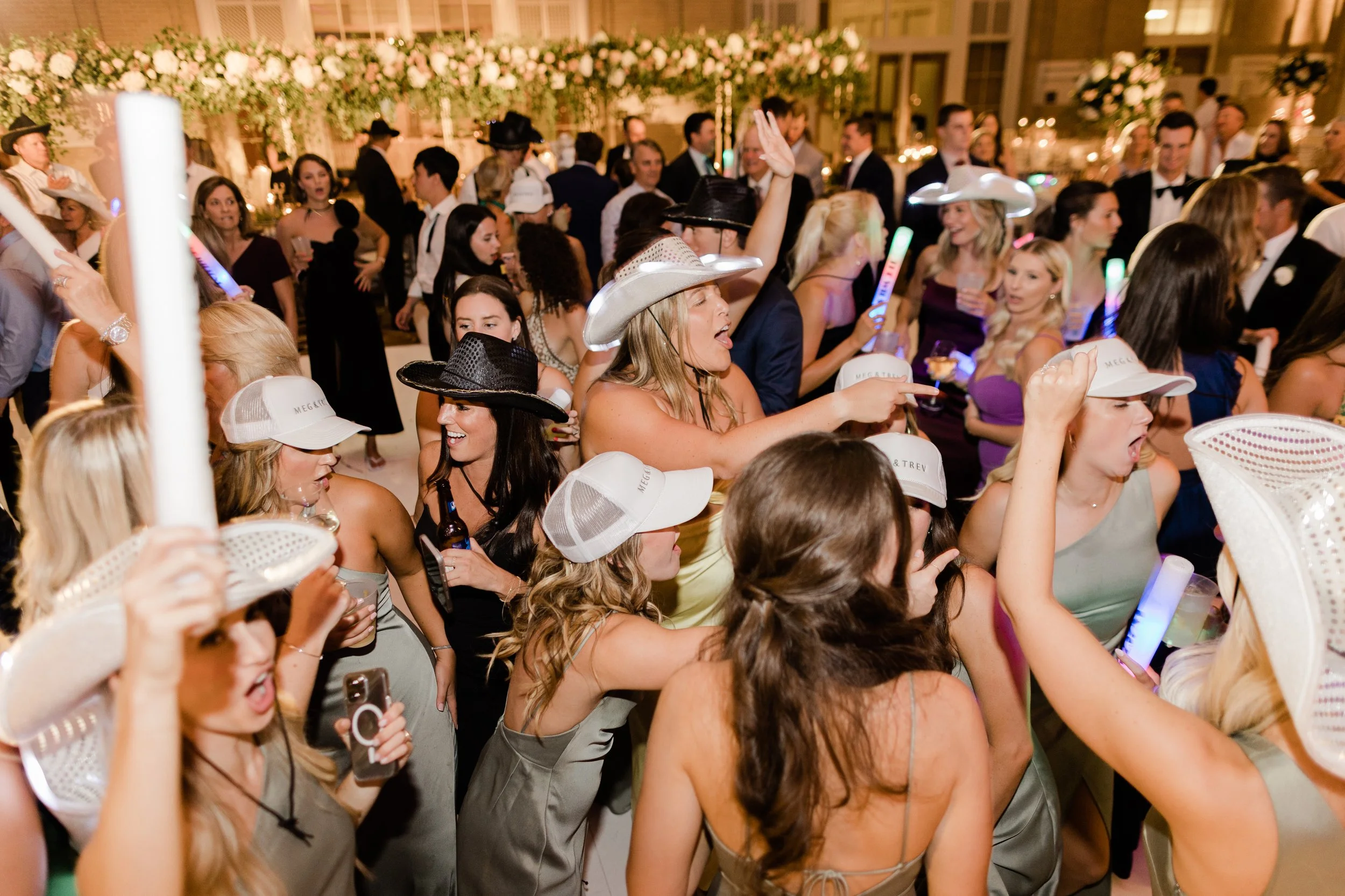 A lively party scene with many women wearing cowboy hats, some holding drinks, and one taking a selfie. In the background, there are more people dressed in formal attire, dancing and enjoying the event.
