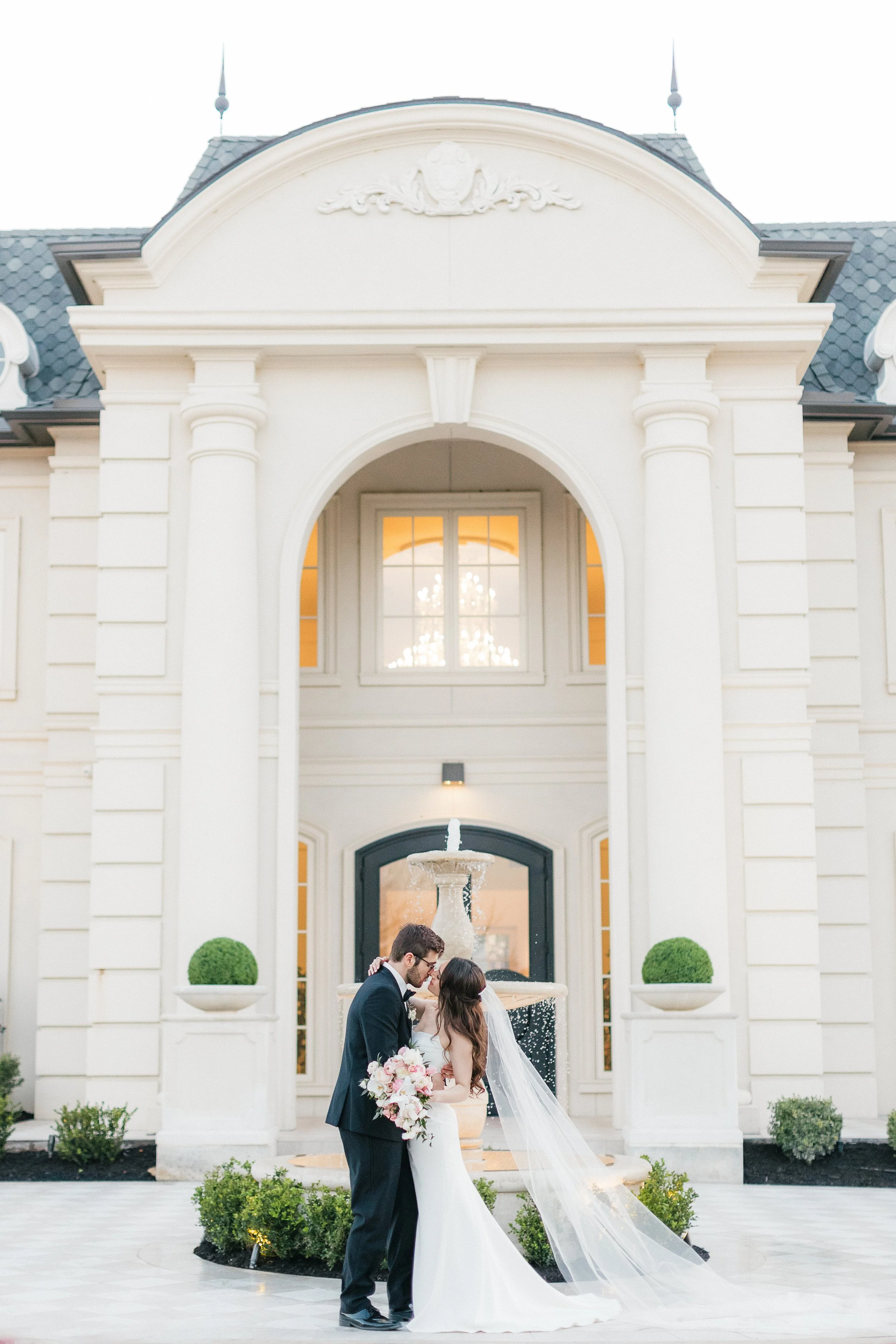 A newlywed couple sharing a kiss in front of a white mansion with a fountain.