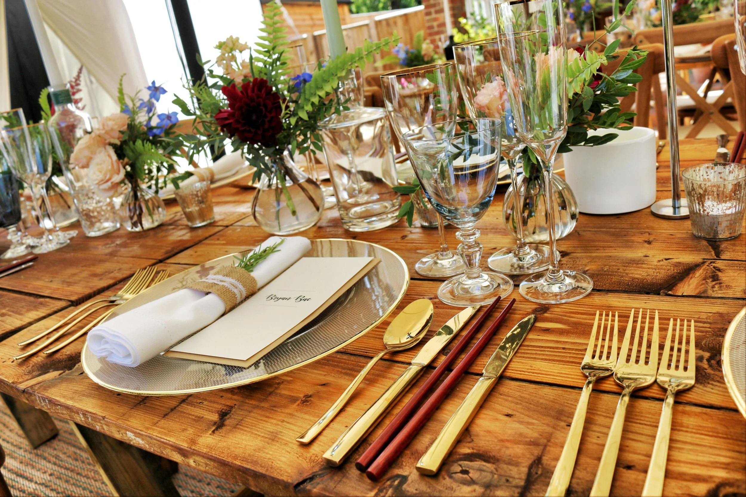 A rustic wooden table set for a formal event with gold and wooden cutlery, glasses, a white napkin with a sprig of herbs, and a menu card, surrounded by floral centerpieces with colorful flowers and greenery.