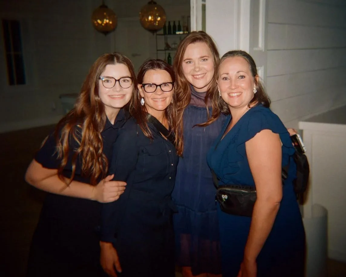 Four women smiling and posing together indoors at a social gathering or event.
