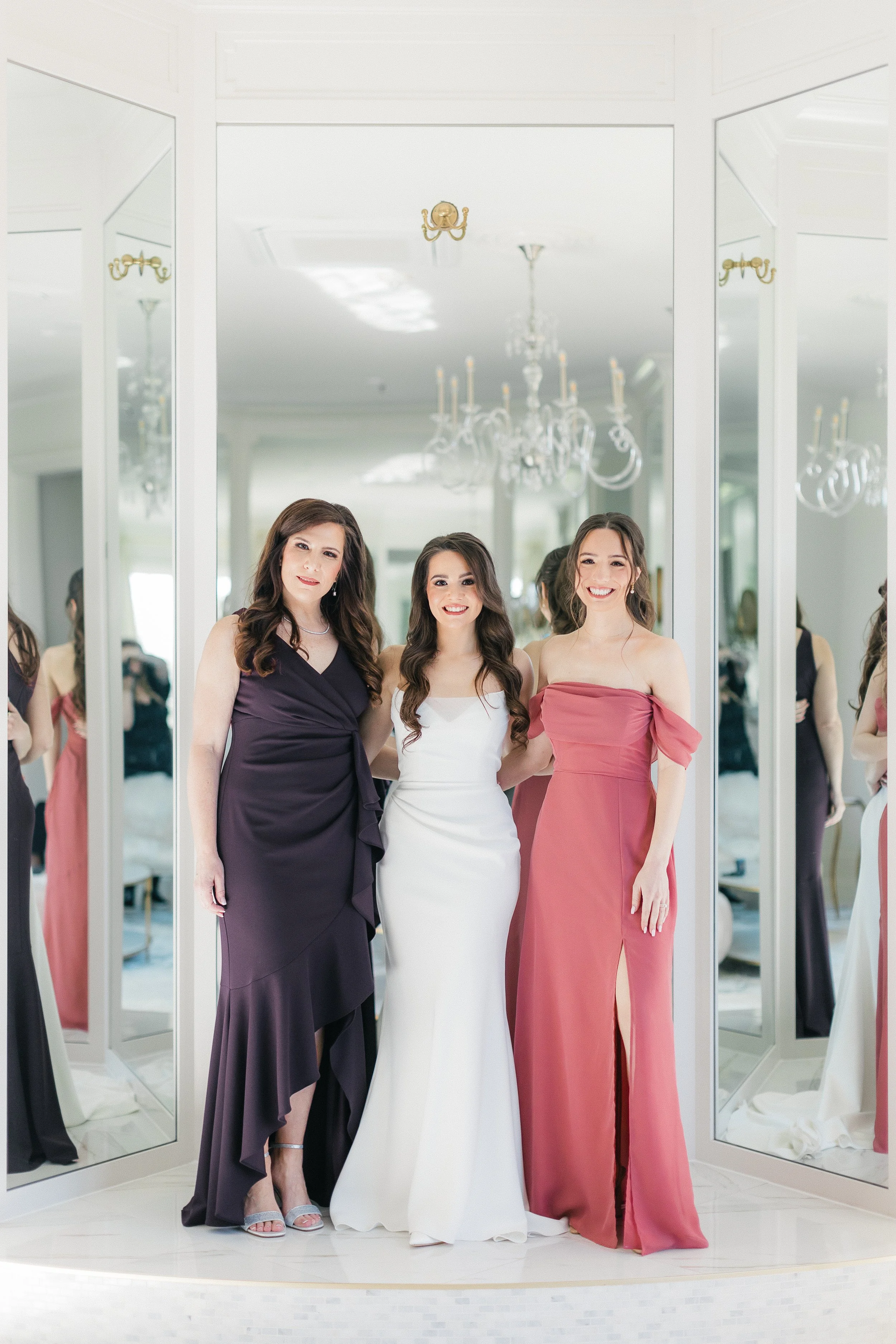 Three women in elegant dresses standing together inside a dressing room with large mirrors and chandeliers.