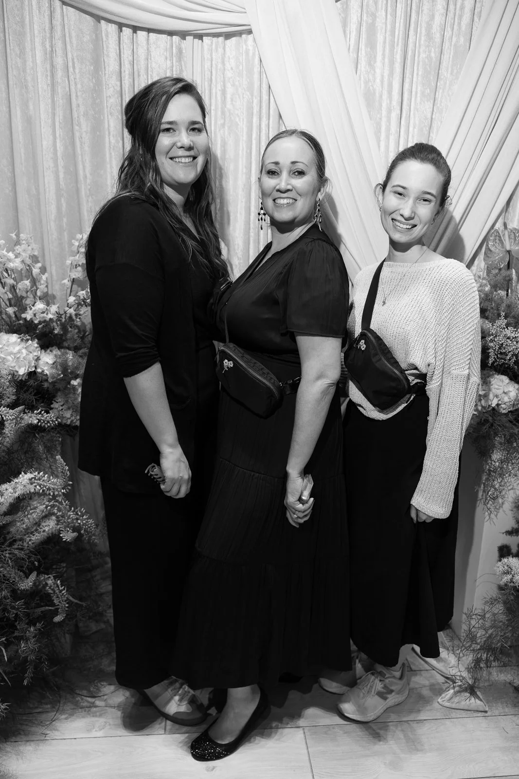 Three women smiling at a photograph in front of floral arrangements and curtains.