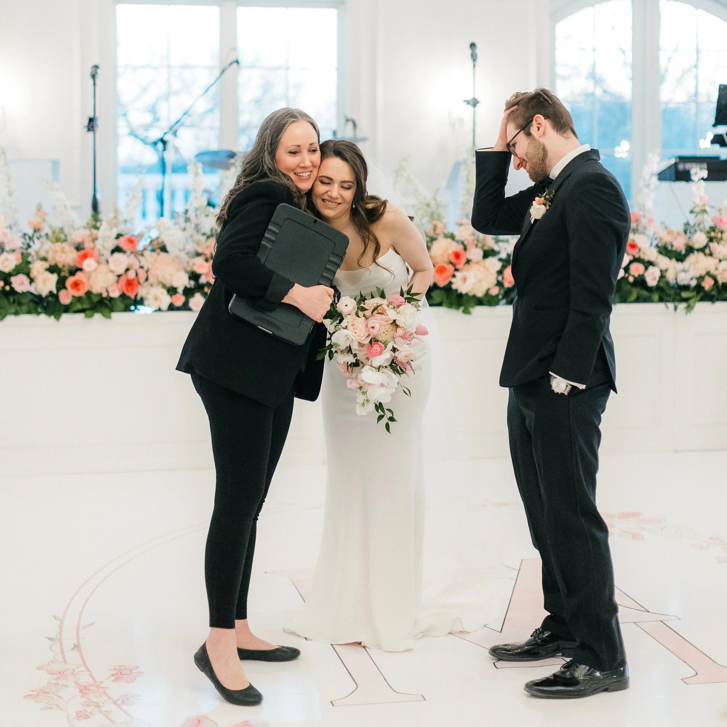 A bride stands with her wedding officiant and groom during a wedding ceremony in a decorated hall with floral arrangements.