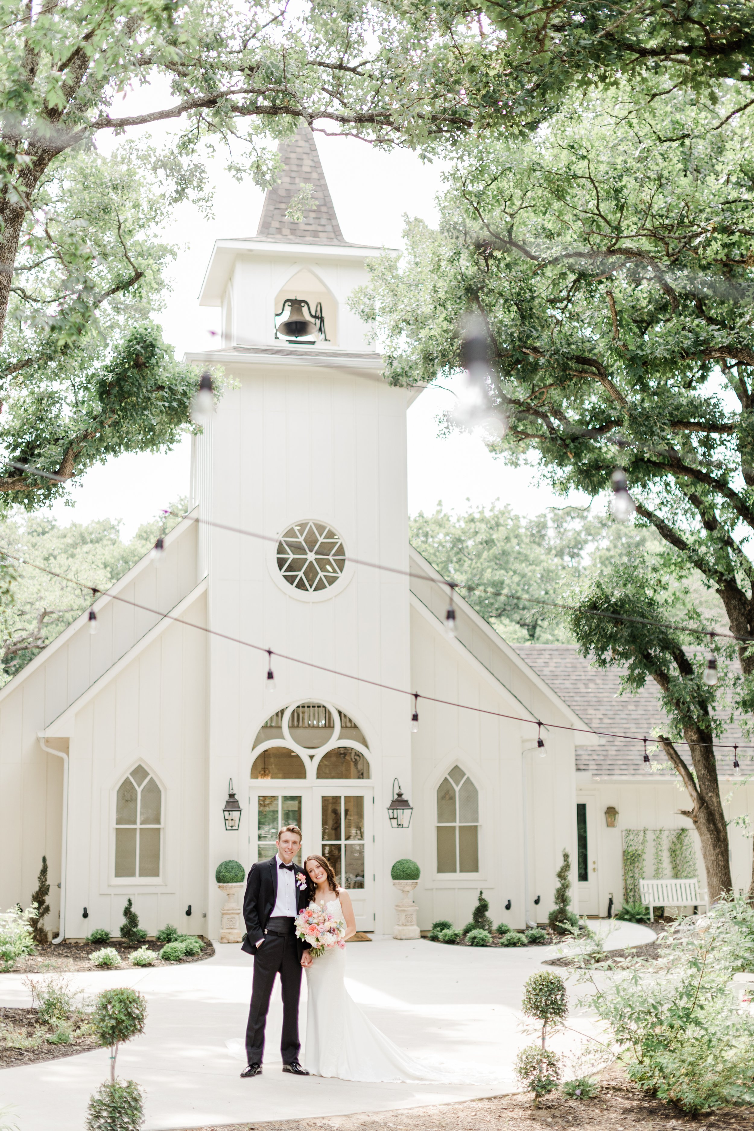 A bride and groom standing in front of a white church with arched windows and a steeple, surrounded by trees and string lights, on their wedding day.