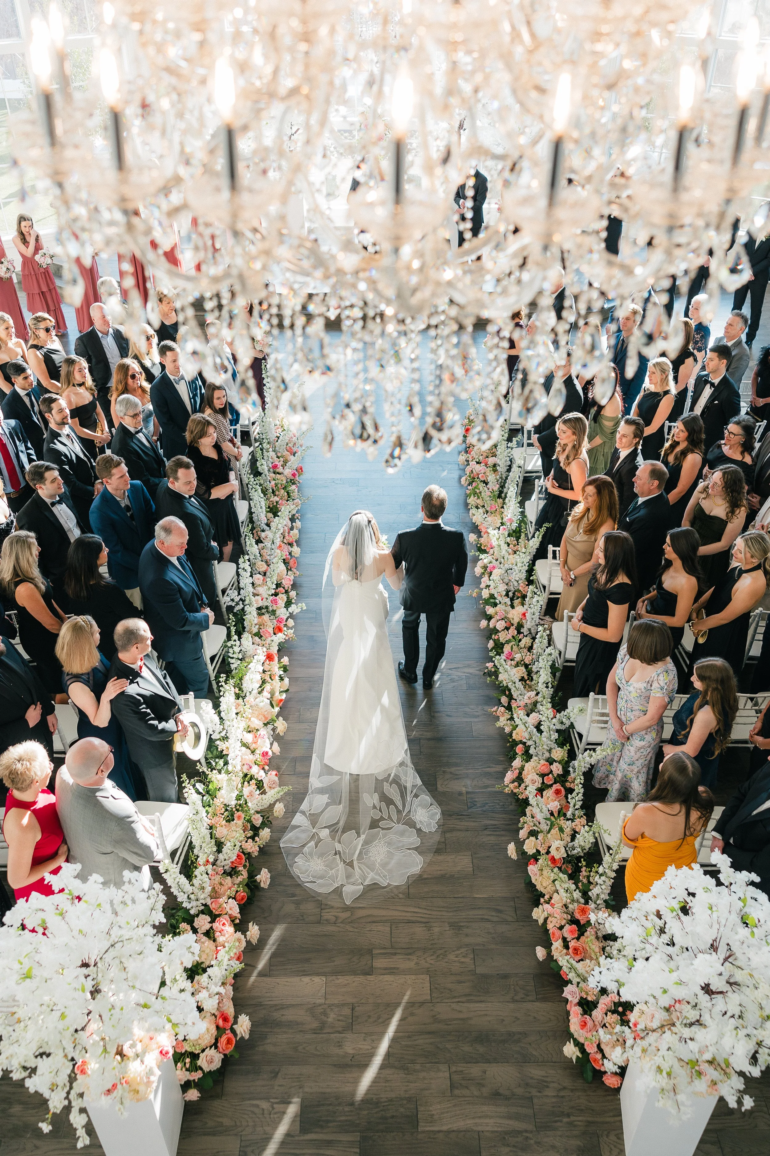 Wedding ceremony with bride and groom walking down the aisle, surrounded by floral arrangements and guests on either side, viewed from above through a chandelier.
