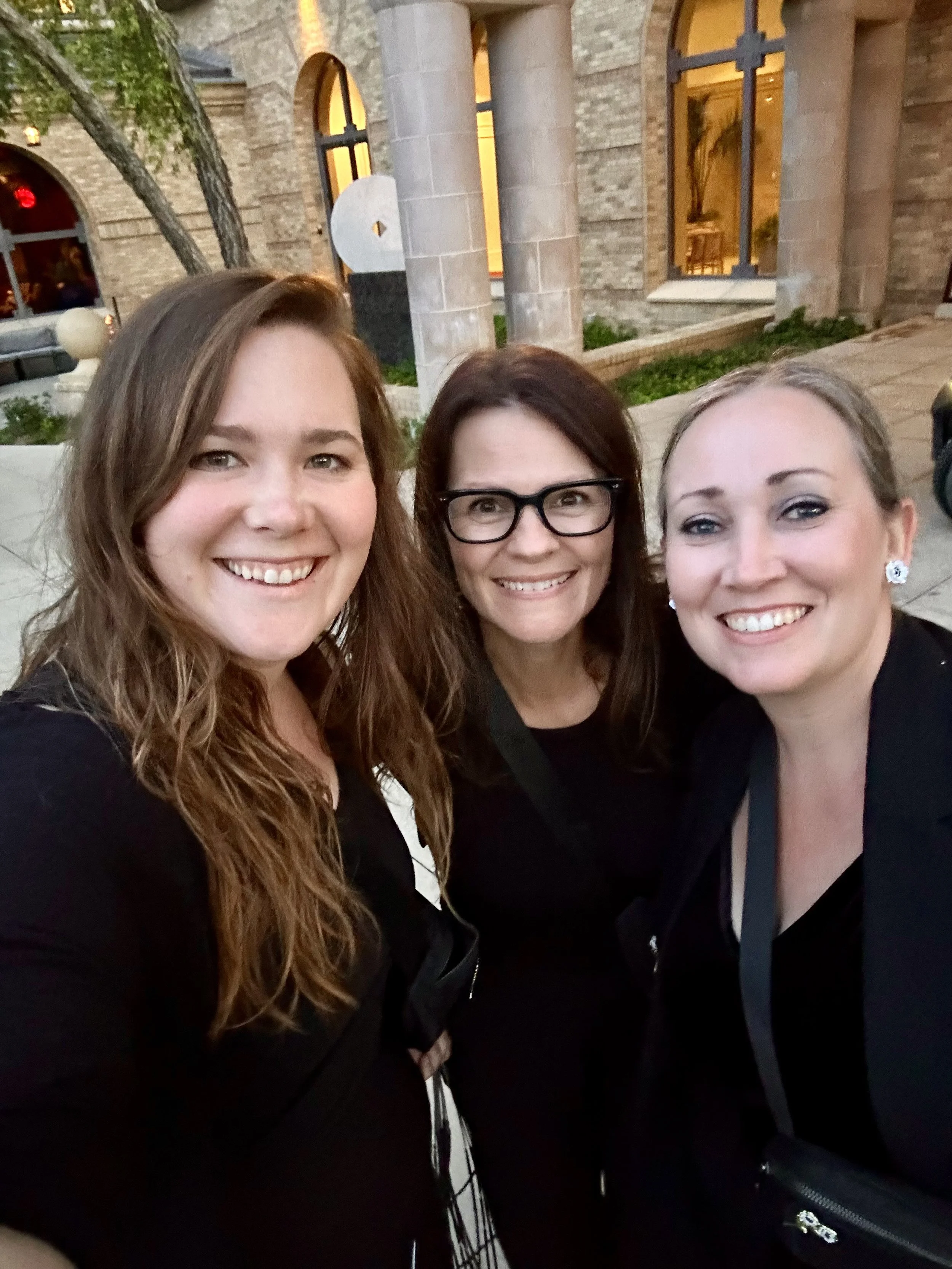 Three women smiling and taking a selfie outdoors in front of a brick building with large windows and some greenery.