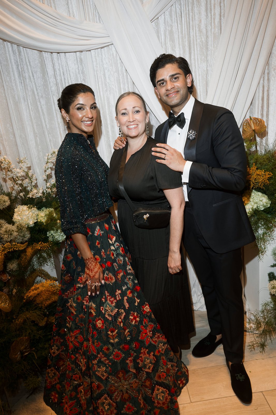 Three people dressed in formal attire standing together at an event, with floral decorations in the background.