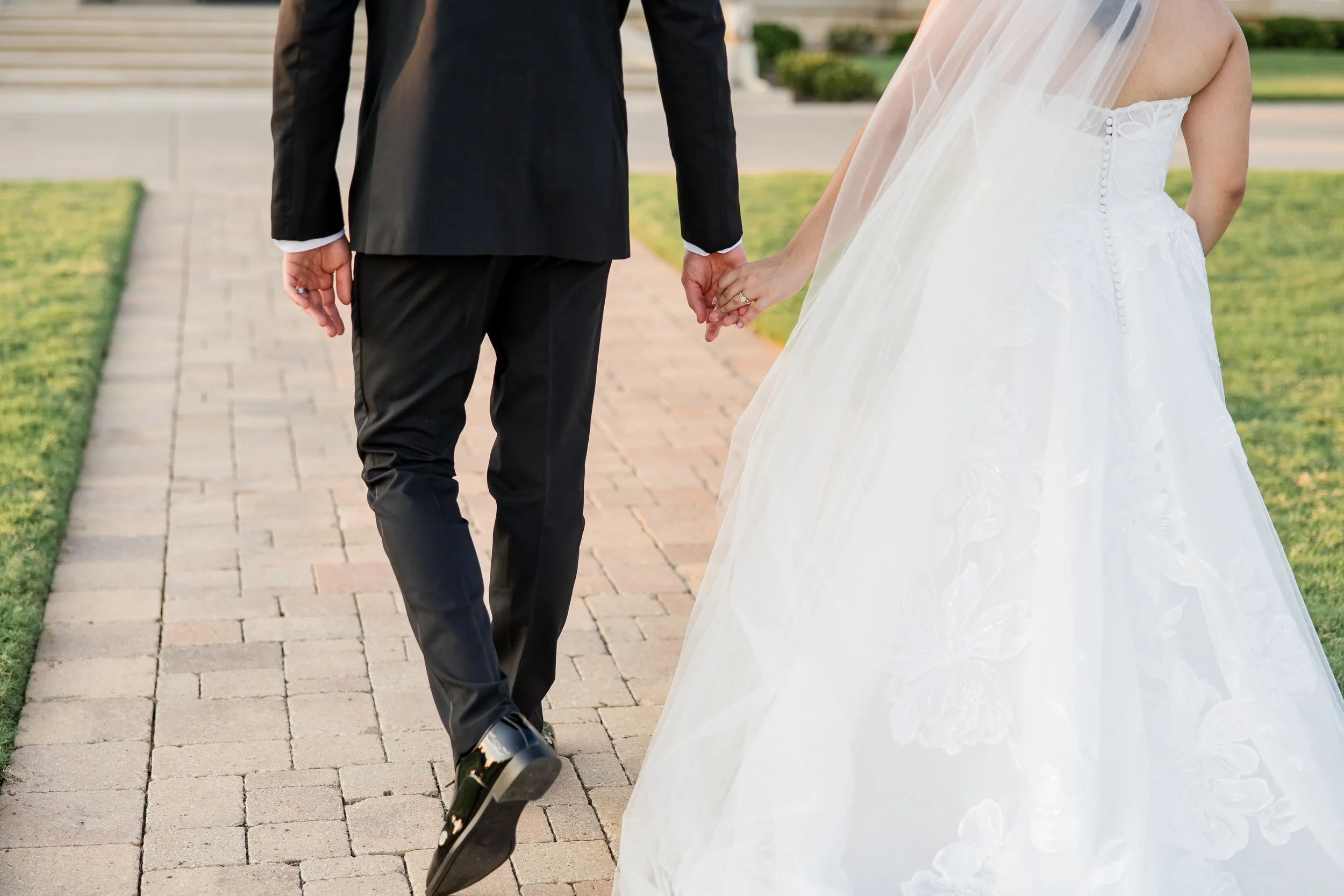 A bride and groom holding hands, walking on a brick pathway outdoors, with the bride in a white wedding gown and veil, and the groom in a black suit.