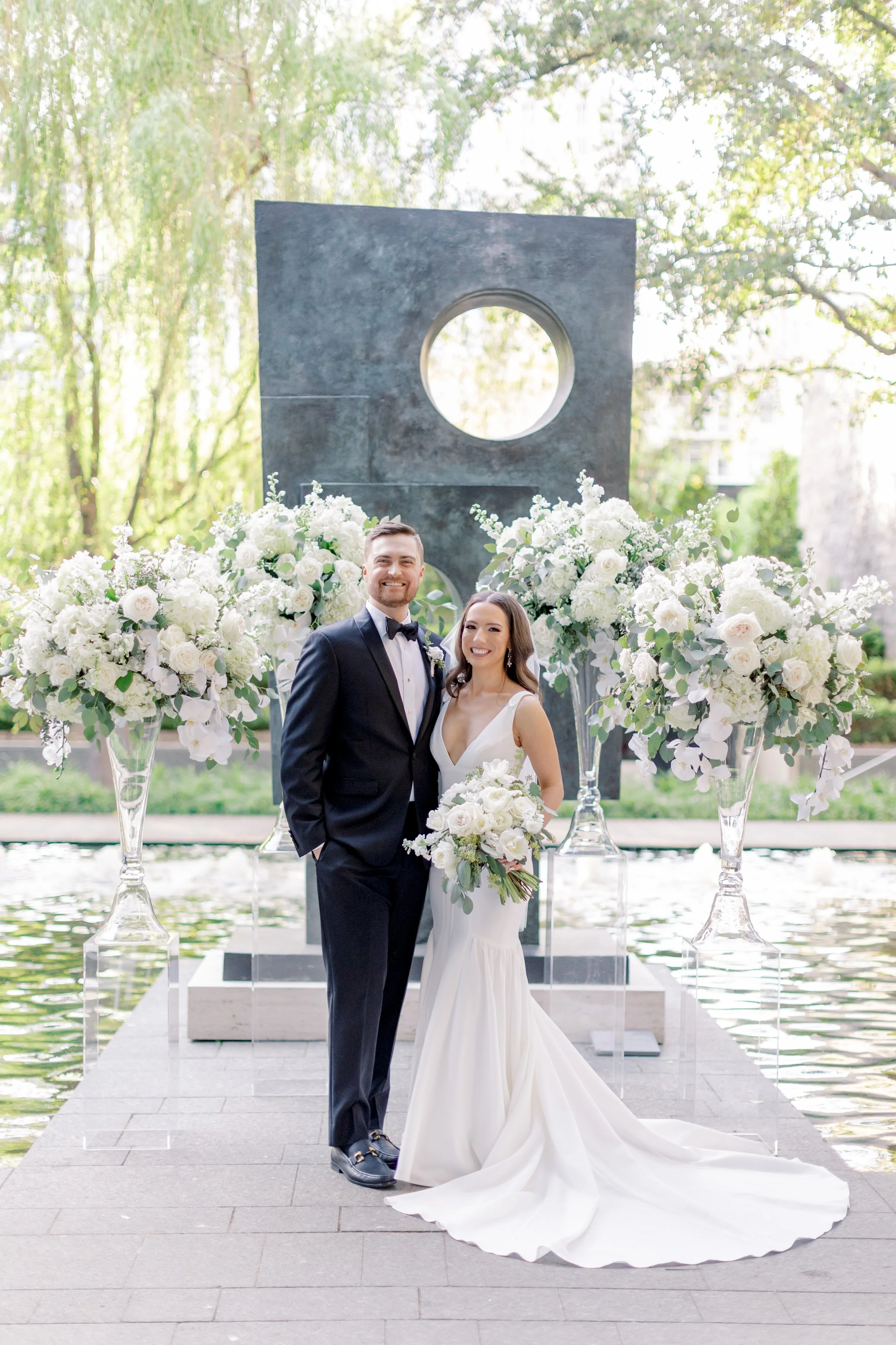 A newlywed couple standing outdoors in front of modern art sculpture, surrounded by large glass vases filled with white flowers, near water and lush green trees.
