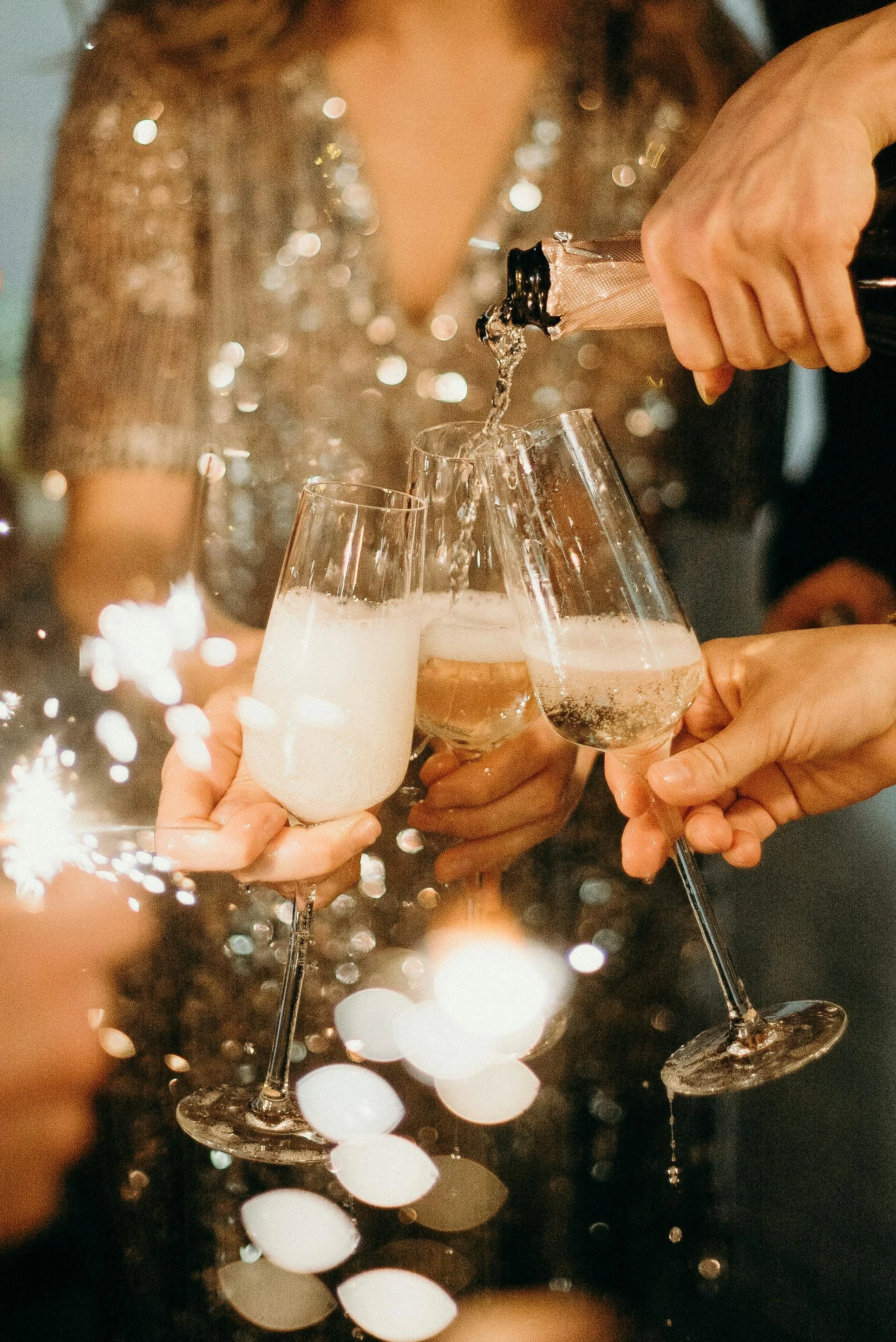 People celebrating with glasses of champagne, one person pouring champagne from a bottle into glass glasses, at a festive event with sparklers and blurred lights in the background.