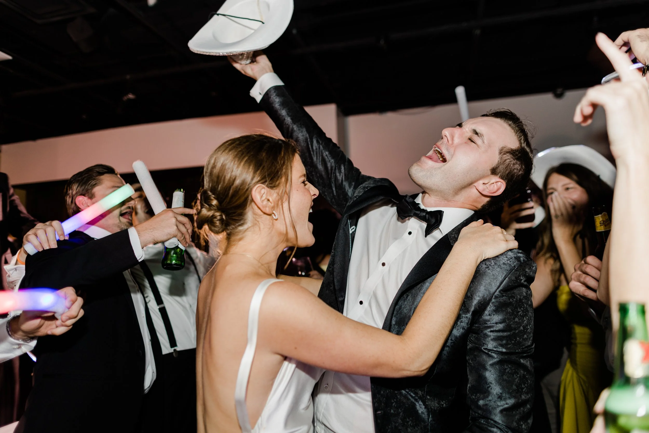 People celebrating at a wedding or party, dancing and laughing, with some holding drinks and glow sticks.
