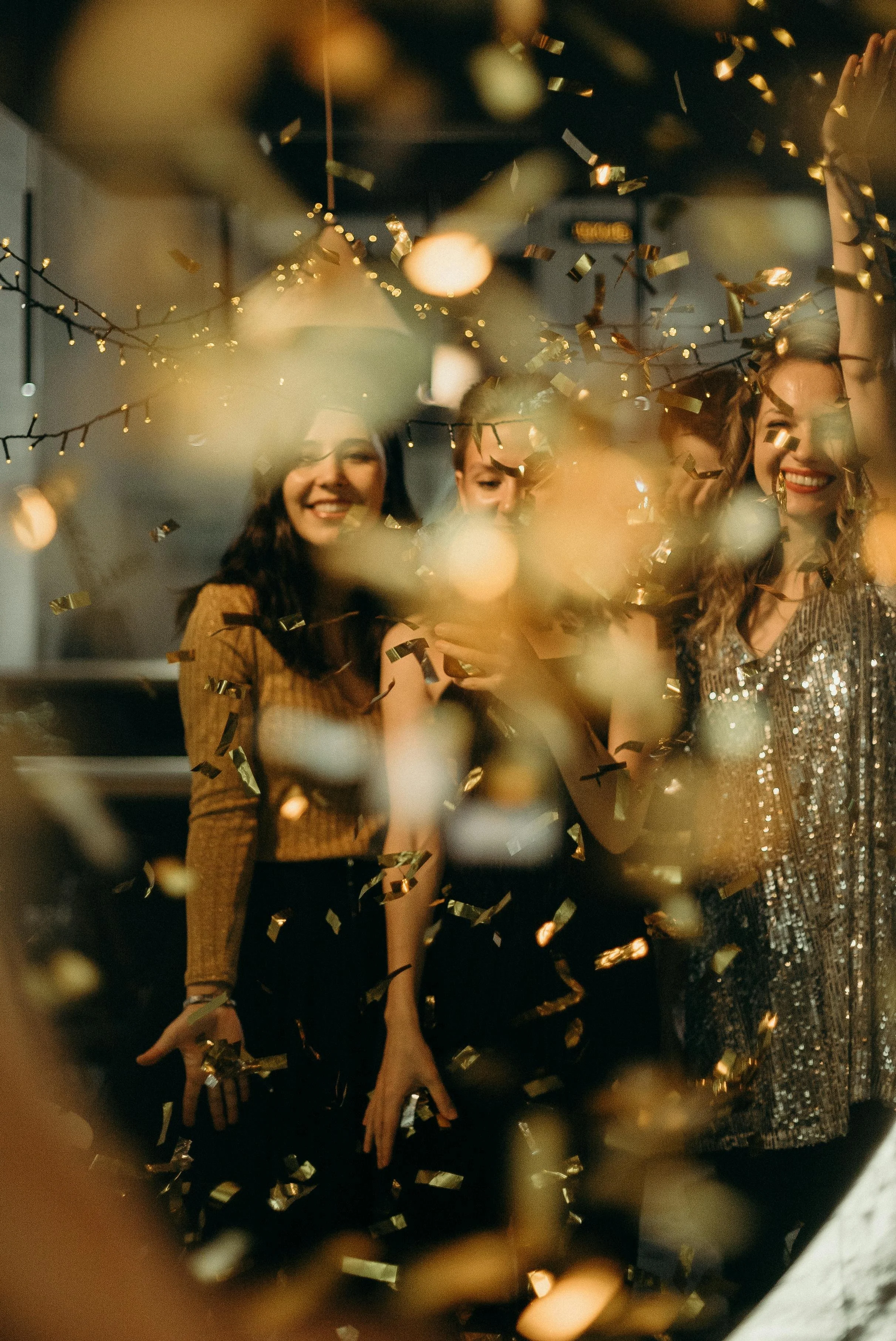 Three women celebrating with confetti, smiling, and wearing party outfits at a festive indoor party.