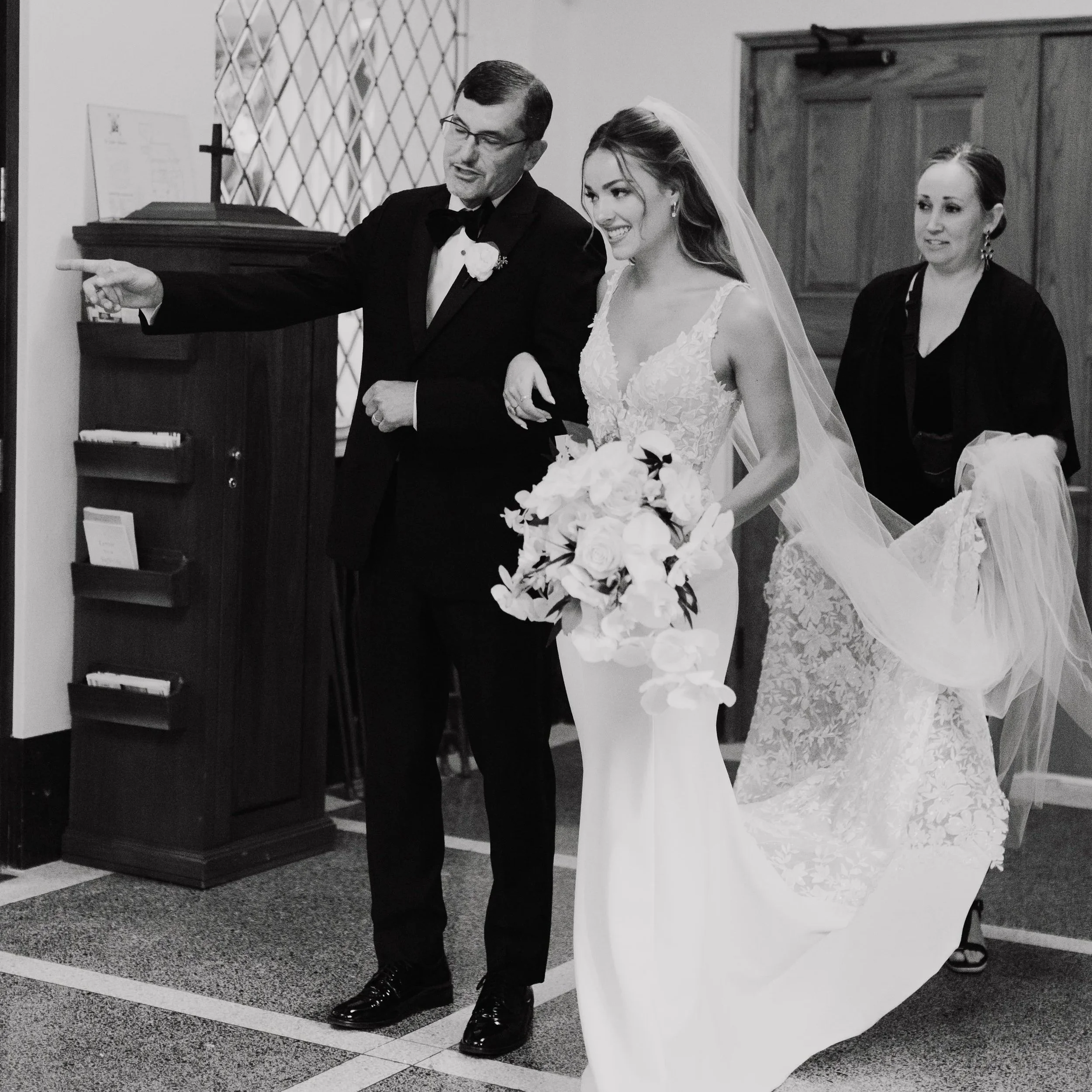 A bride in a white lace wedding gown and veil holding a bouquet of flowers walks down the aisle with a man in a tuxedo. An officiant stands behind them. They are indoors, with decorative wooden panels and a piano in the background.