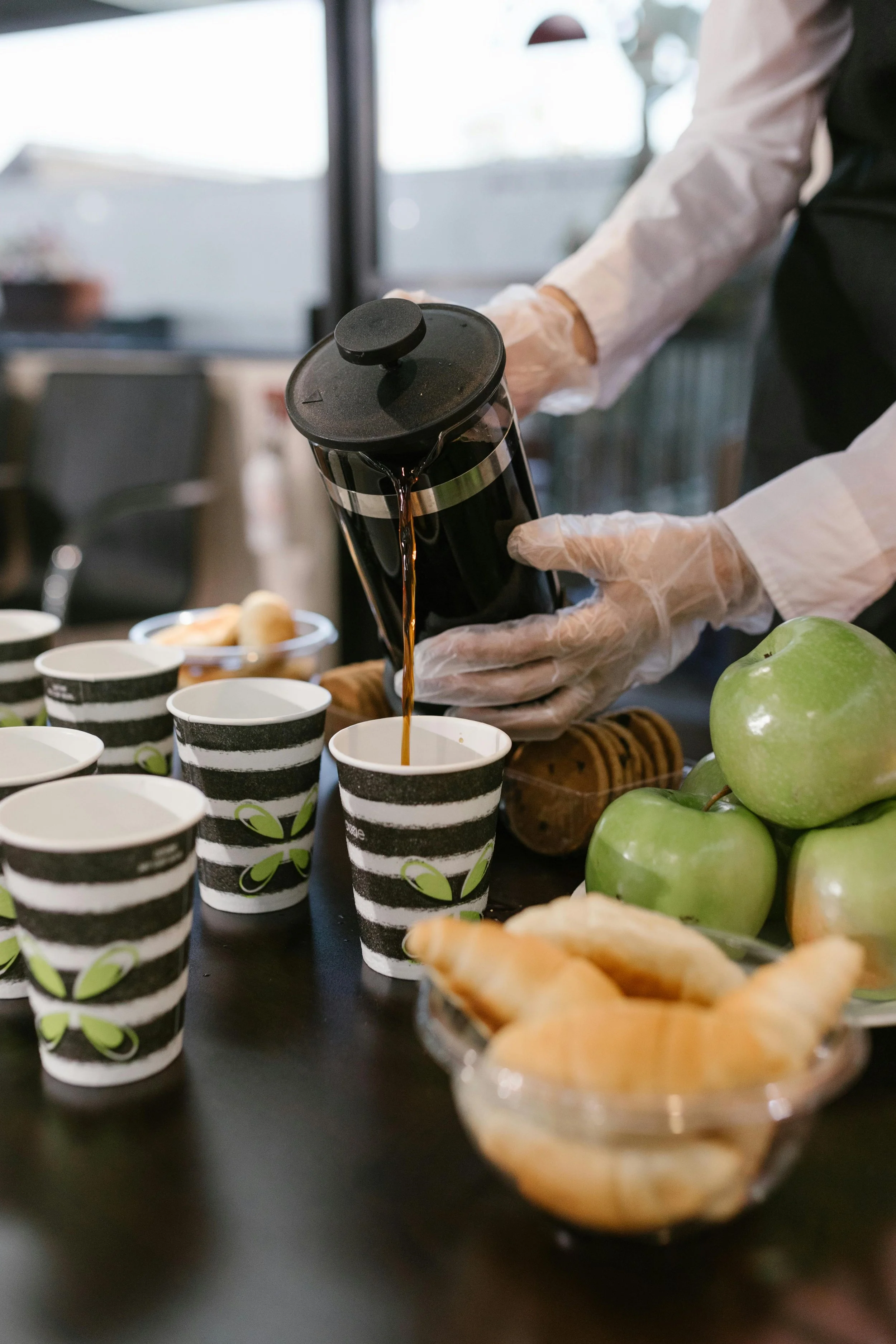 Person pouring coffee into striped paper cups at a table with apples, croissants, and cookies, in a bright, modern setting.