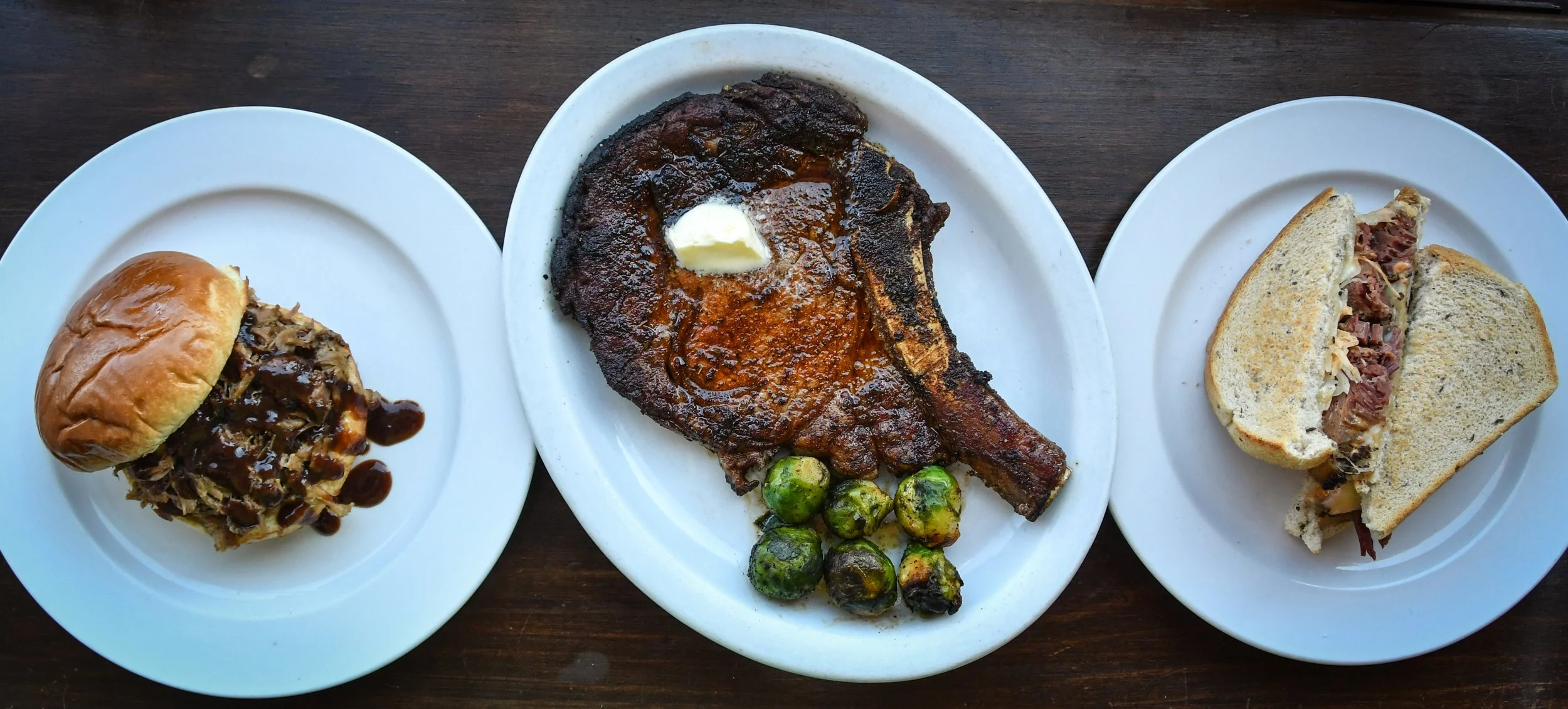Plate with grilled pork chop topped with butter, roasted Brussels sprouts, plate with barbecue slider with beef and barbecue sauce, and a chopped beef sandwich with bun.