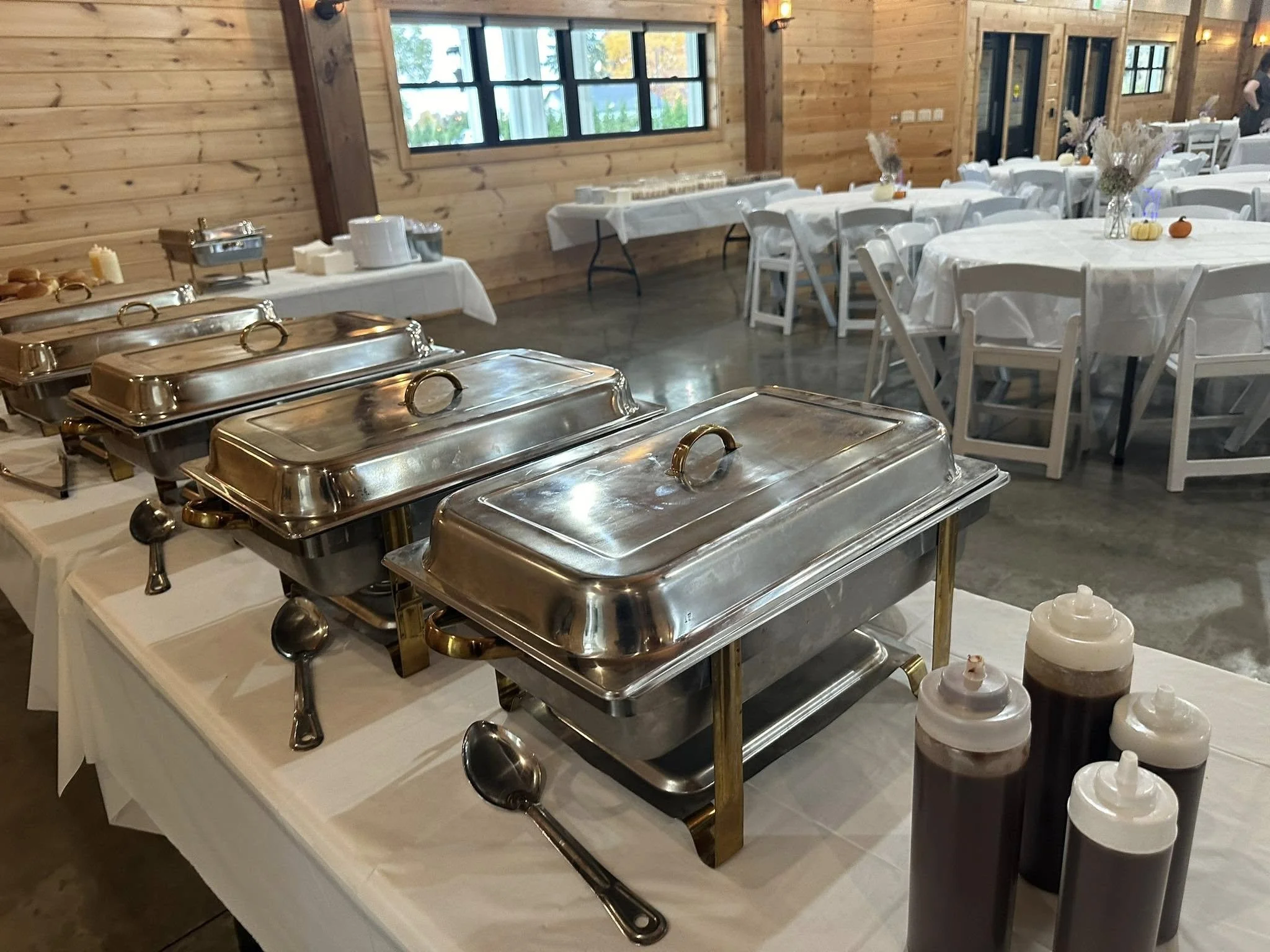 Buffet setup with several silver chafing dishes on a table, with attached serving spoons, in a rustic event hall with white tablecloths, white chairs, and decoration pumpkins on the tables.