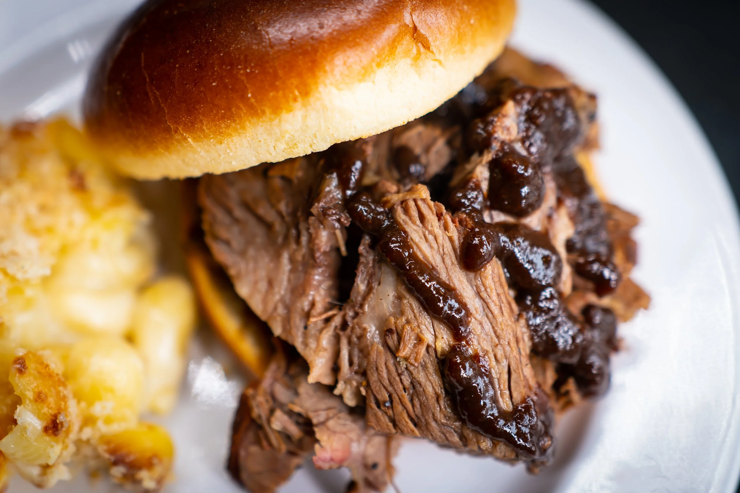 Close-up of a pulled pork sandwich with barbecue sauce on a bun, served with macaroni and cheese on a white plate.