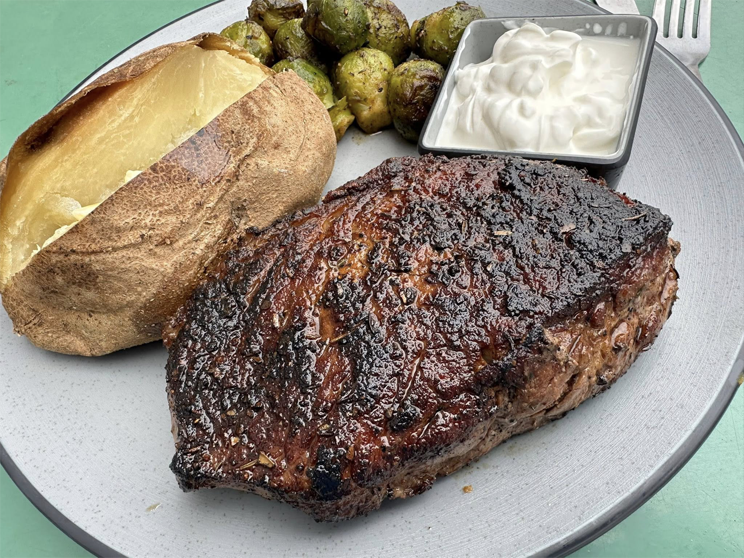 A plate of steak, baked potato with butter, roasted Brussels sprouts, and a small bowl of sour cream.