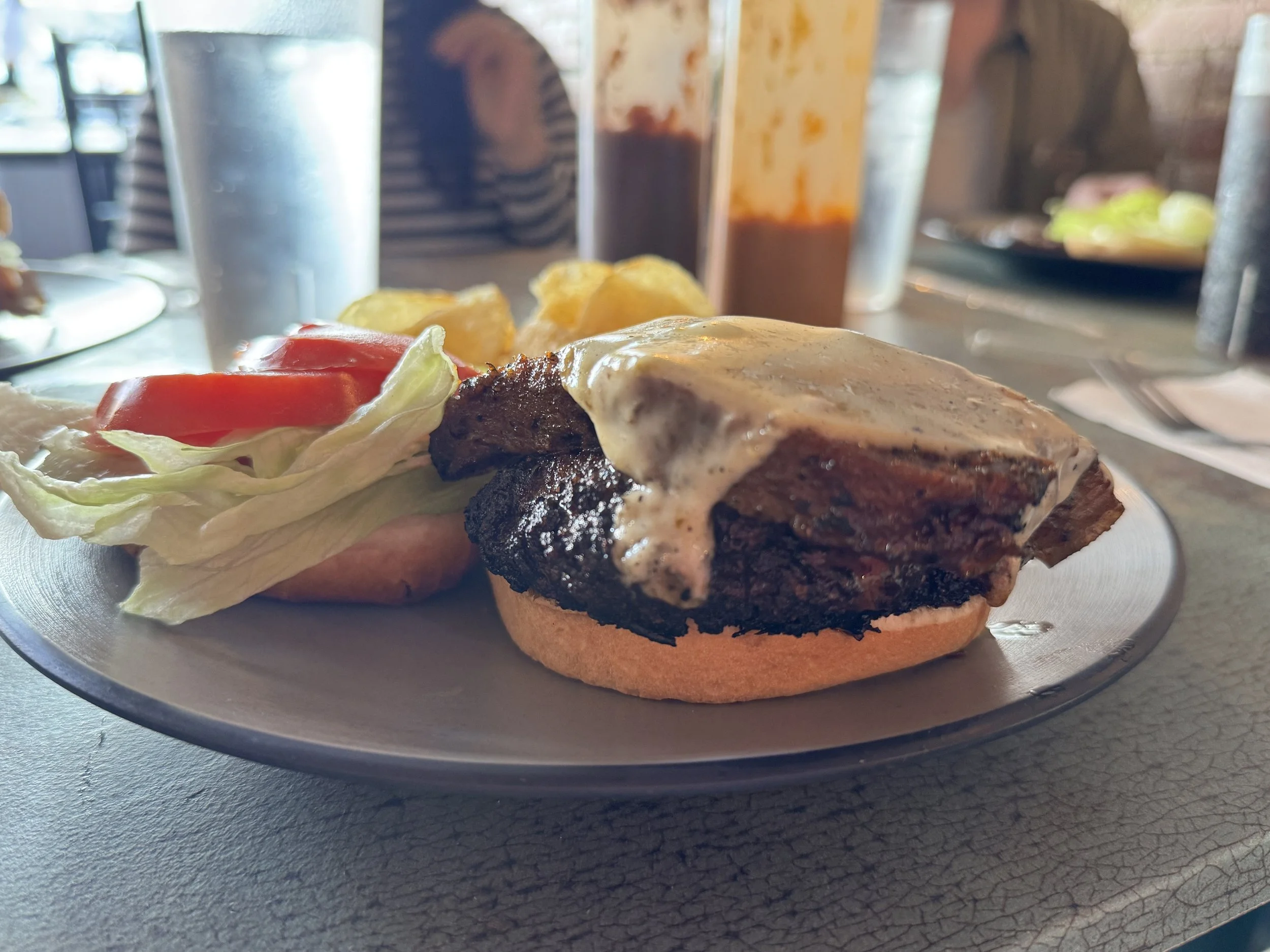 Hamburger with melted cheese, lettuce, tomato slices, onion, served with potato wedges and condiments in the background.