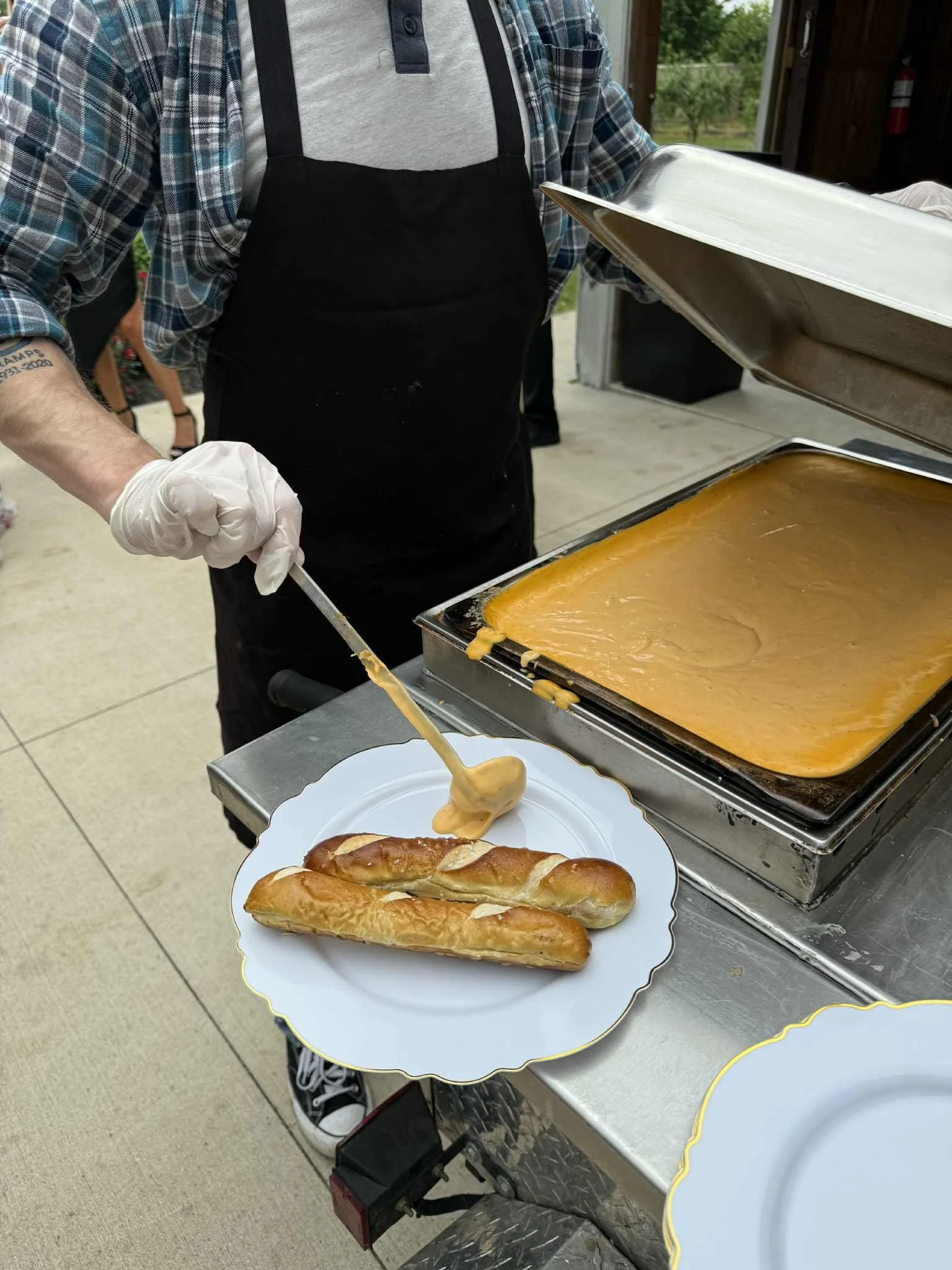 A person wearing a black apron and plaid shirt preparing hot cheese sandwiches with melted cheese using a spatula on a silver tray, with a white plate holding breadsticks nearby.