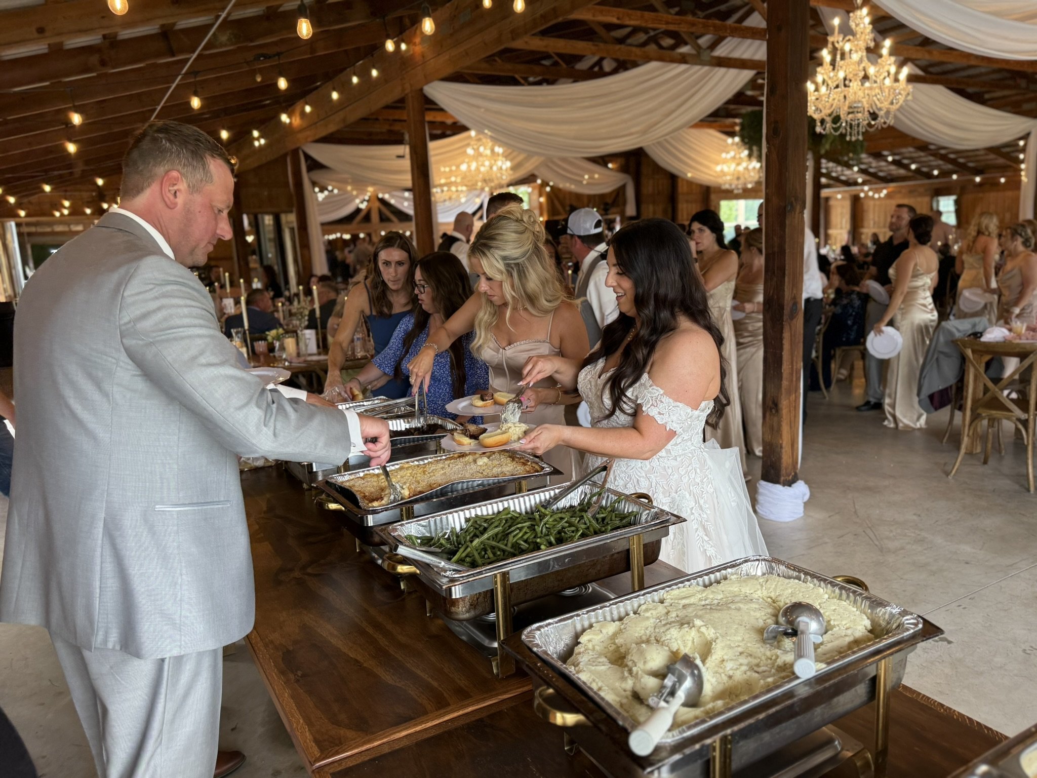 People in wedding attire at a buffet table with food at a wedding reception, decorated with string lights, chandeliers, and draped fabric in a wooden barn venue.