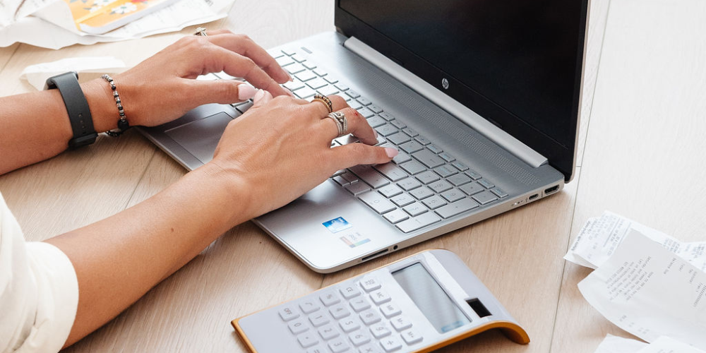 Person working on a laptop with a calculator and receipts on a wooden desk.