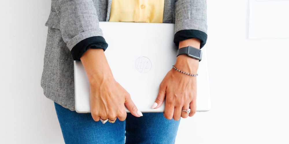 Person holding a closed white HP laptop with both hands, wearing a smartwatch and jewelry, standing against a plain white wall.