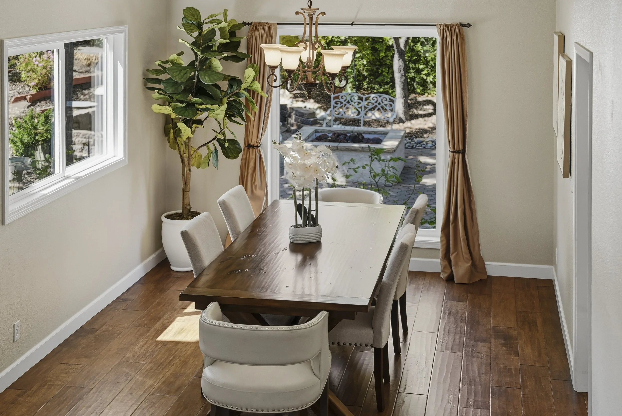 A dining room with a wooden table, six upholstered chairs, a large potted fiddle leaf fig plant, a chandelier with five lights, and an open sliding glass door leading to an outdoor patio with a stone fire pit, a white metal bench, and trees in the background.