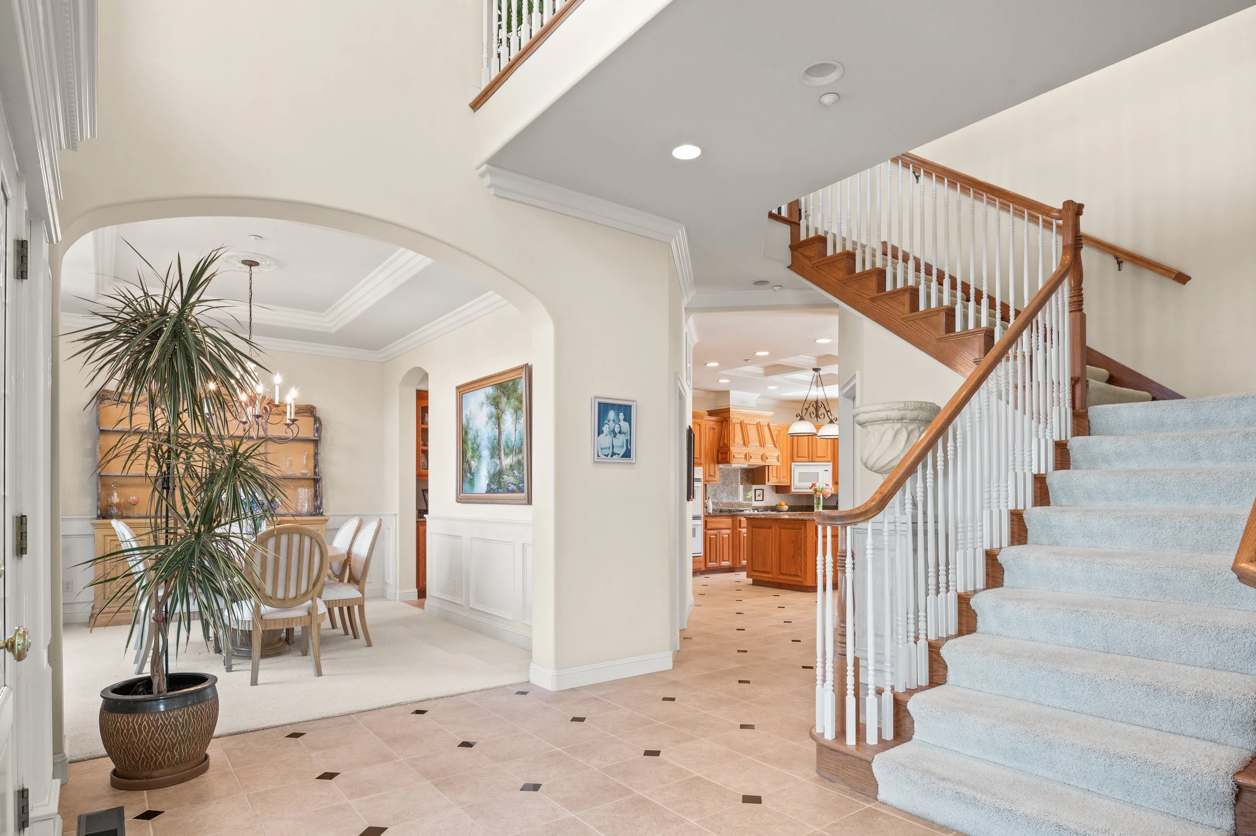 Interior of a house entrance with tiled floor, staircase with wooden handrail and white spindles, dining area with chandelier, and view into the kitchen with wooden cabinets and a centerpiece of flowers.