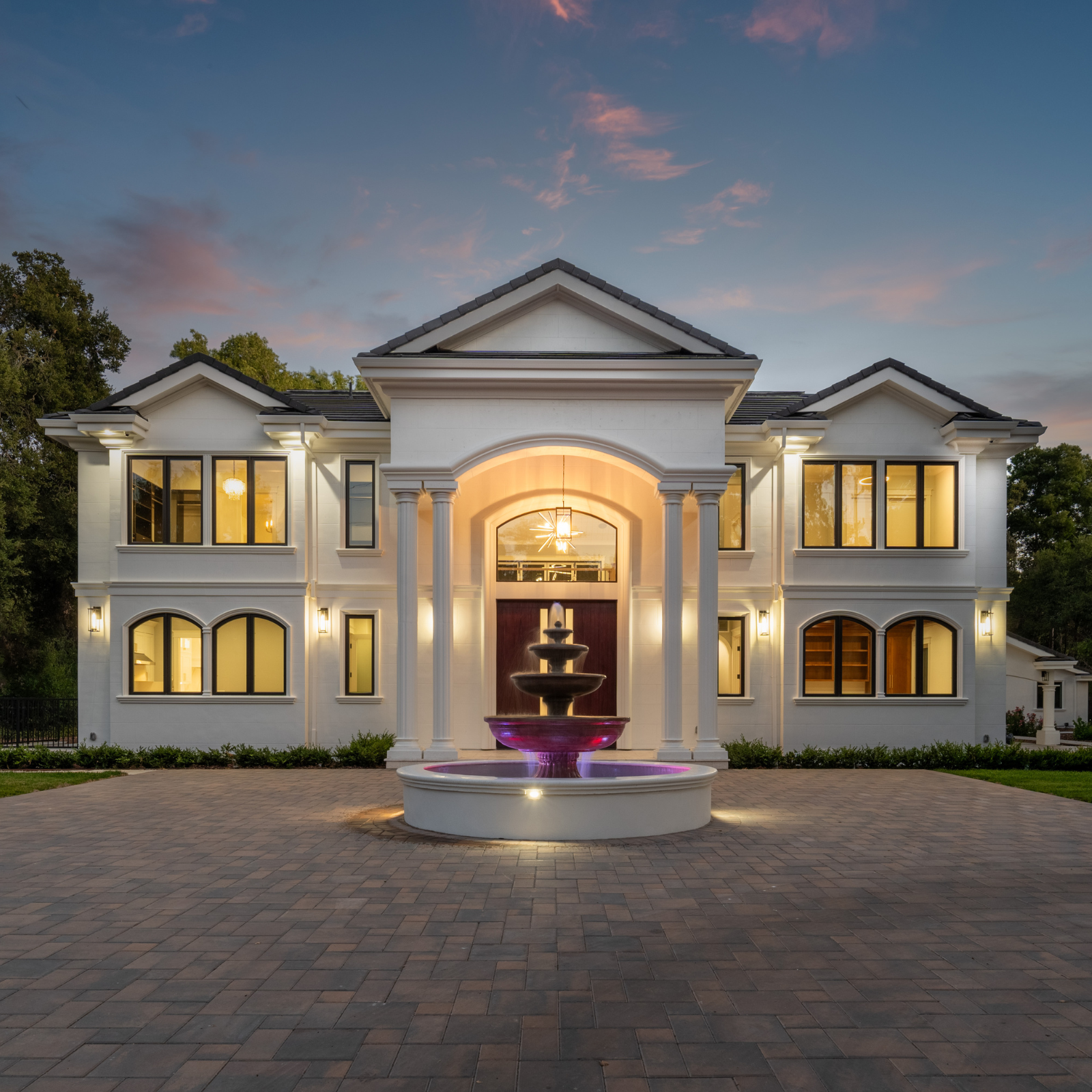A large luxurious white house with multiple windows, columns, and a fountain in the front yard at dusk. The house and fountain are illuminated, with a soft sunset sky overhead.