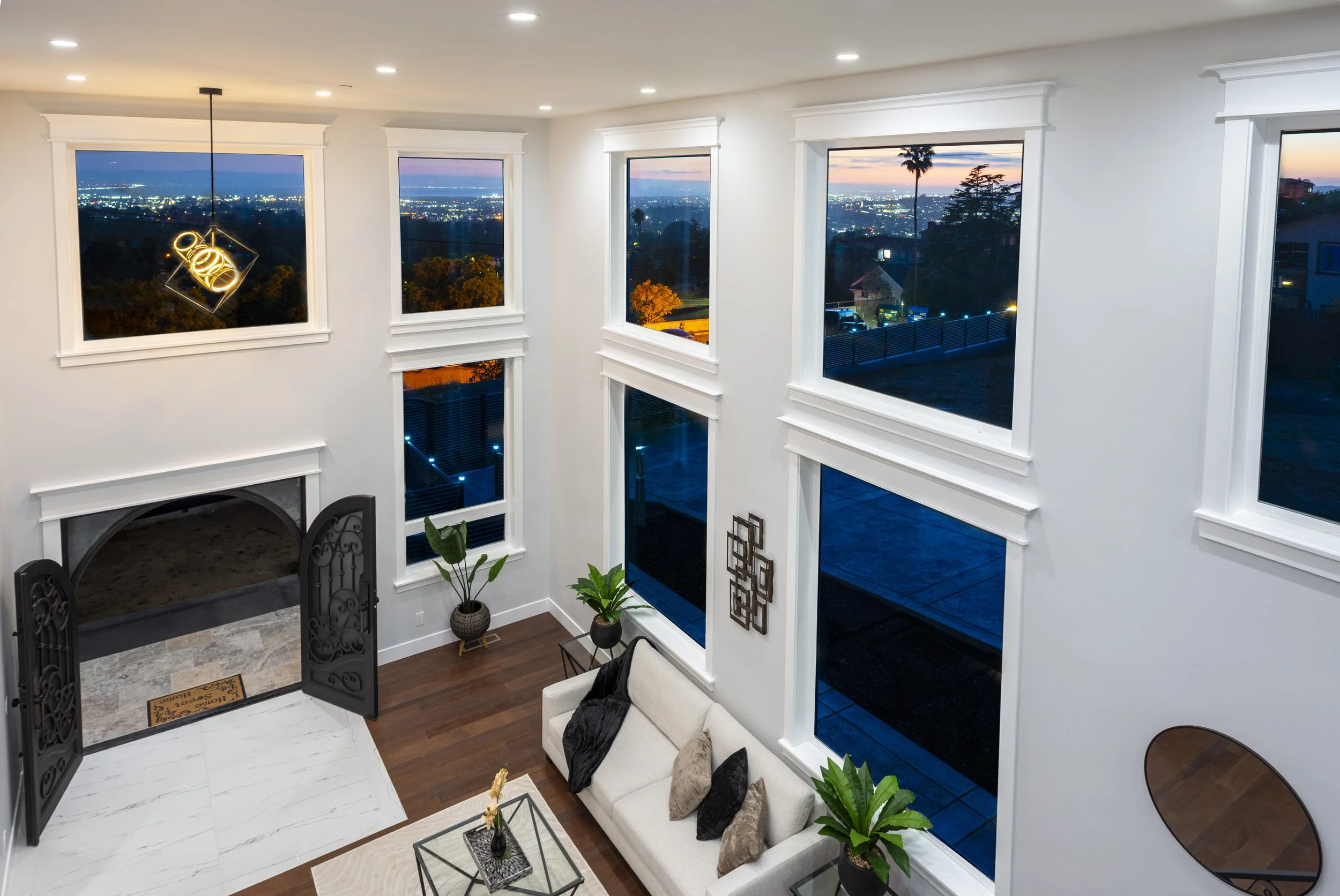 Interior of a modern living room with high ceilings, large windows with white trim showcasing a cityscape at sunset, a cream-colored sofa with pillows, a black armchair, small tables with plants, and a view of the sky turning pink and purple.