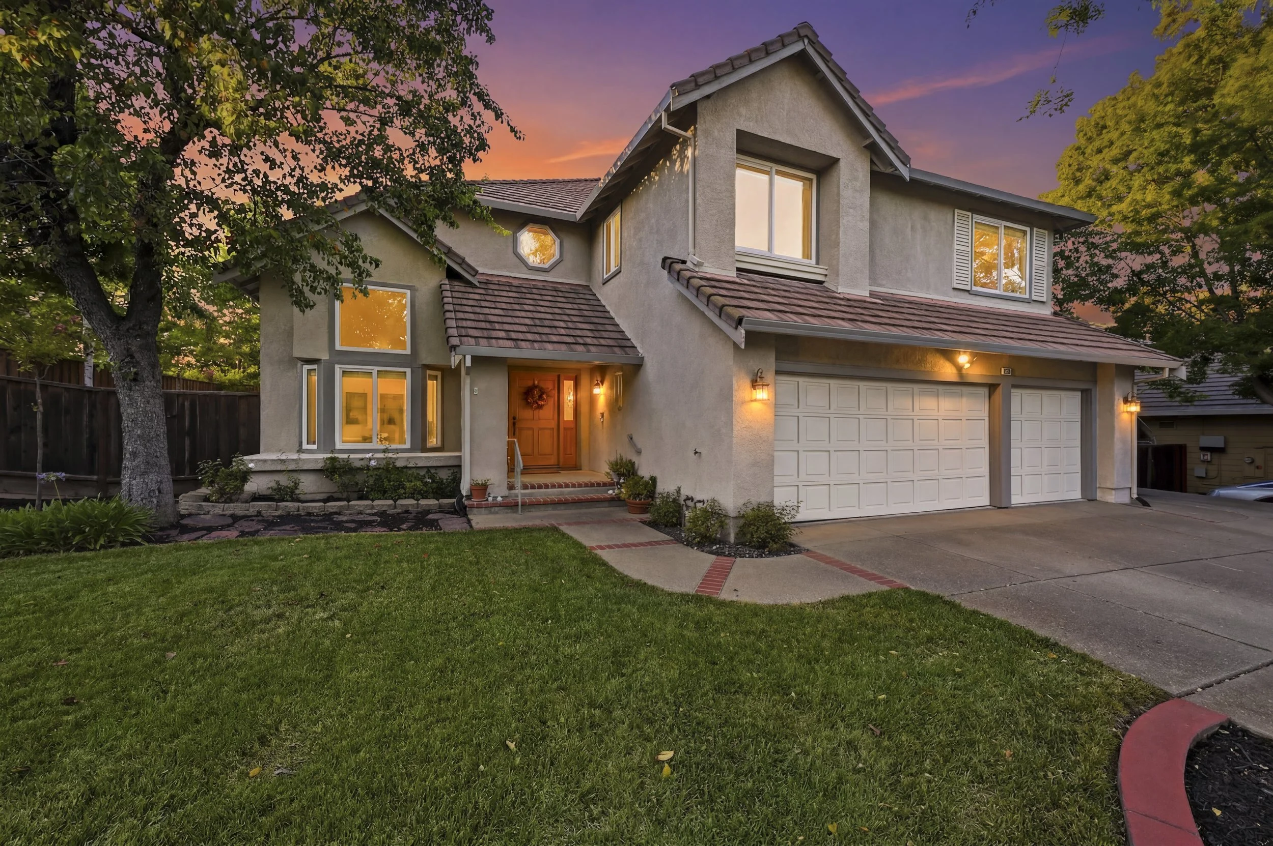 Front view of a two-story house with a manicured lawn and trees, illuminated by outdoor lights at dusk, with a colorful sky in the background.