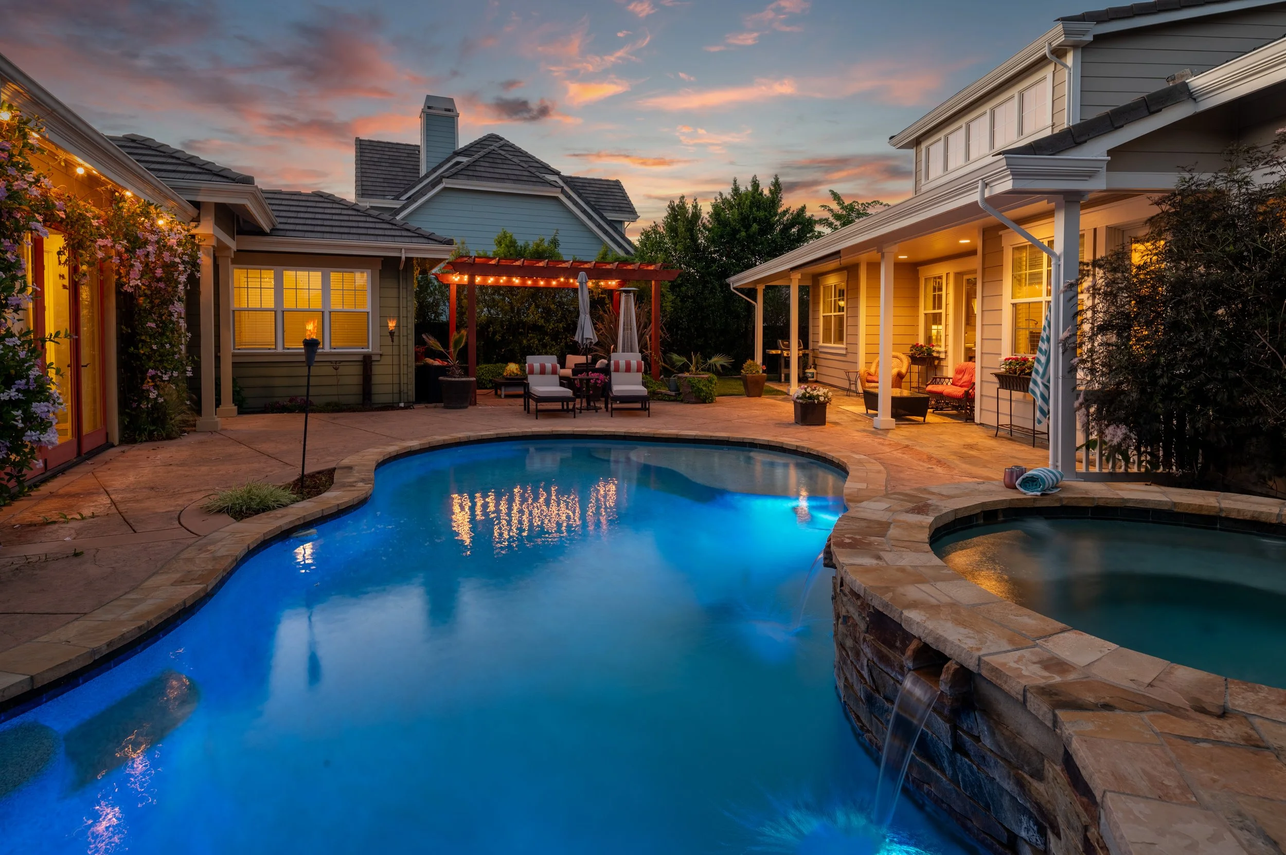 A backyard with a swimming pool under a colorful sunset sky, surrounded by houses with lit windows, outdoor furniture, and decorative plants.