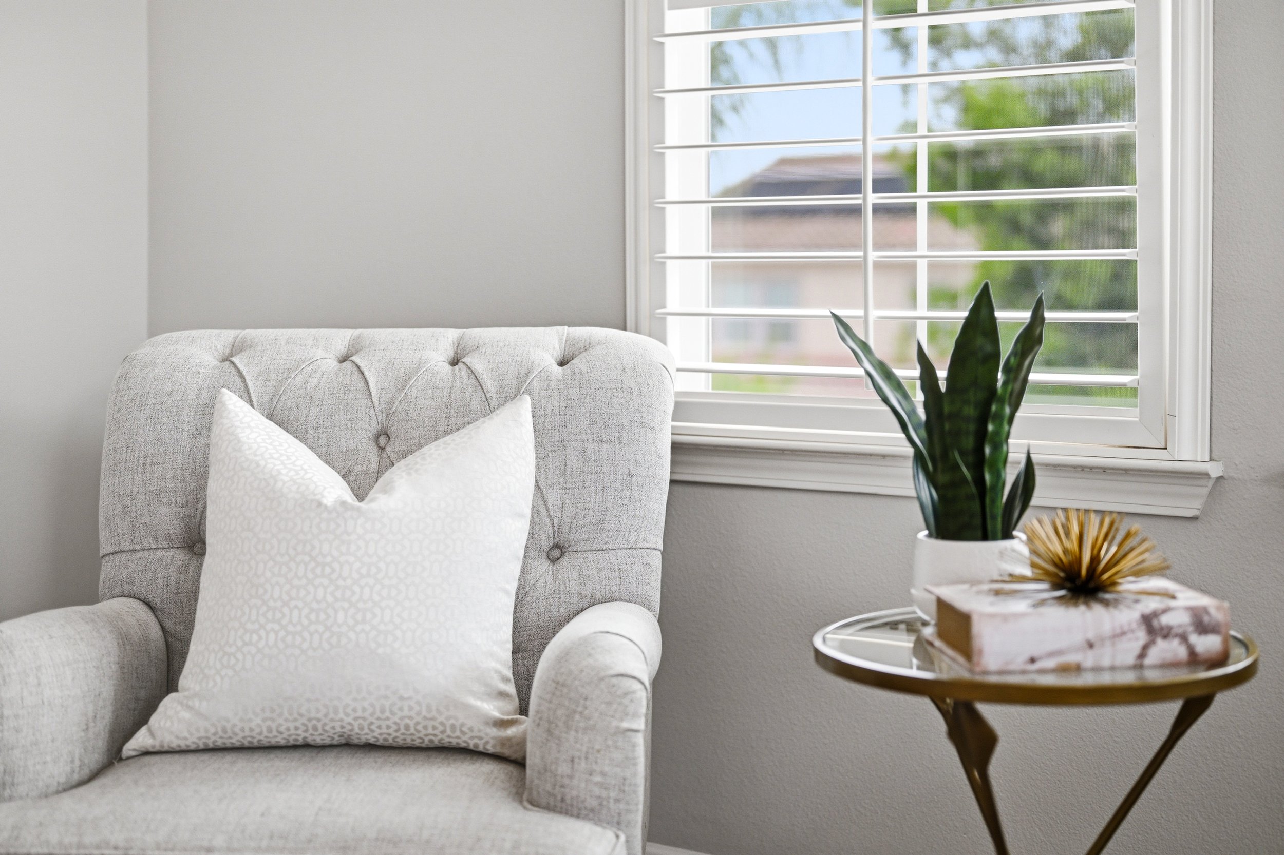 A cozy living room corner featuring a light gray tufted armchair with white patterned pillows, a large window with white shutters letting in natural light, a green potted plant on a side table, and decorative gold-colored accents on the table.