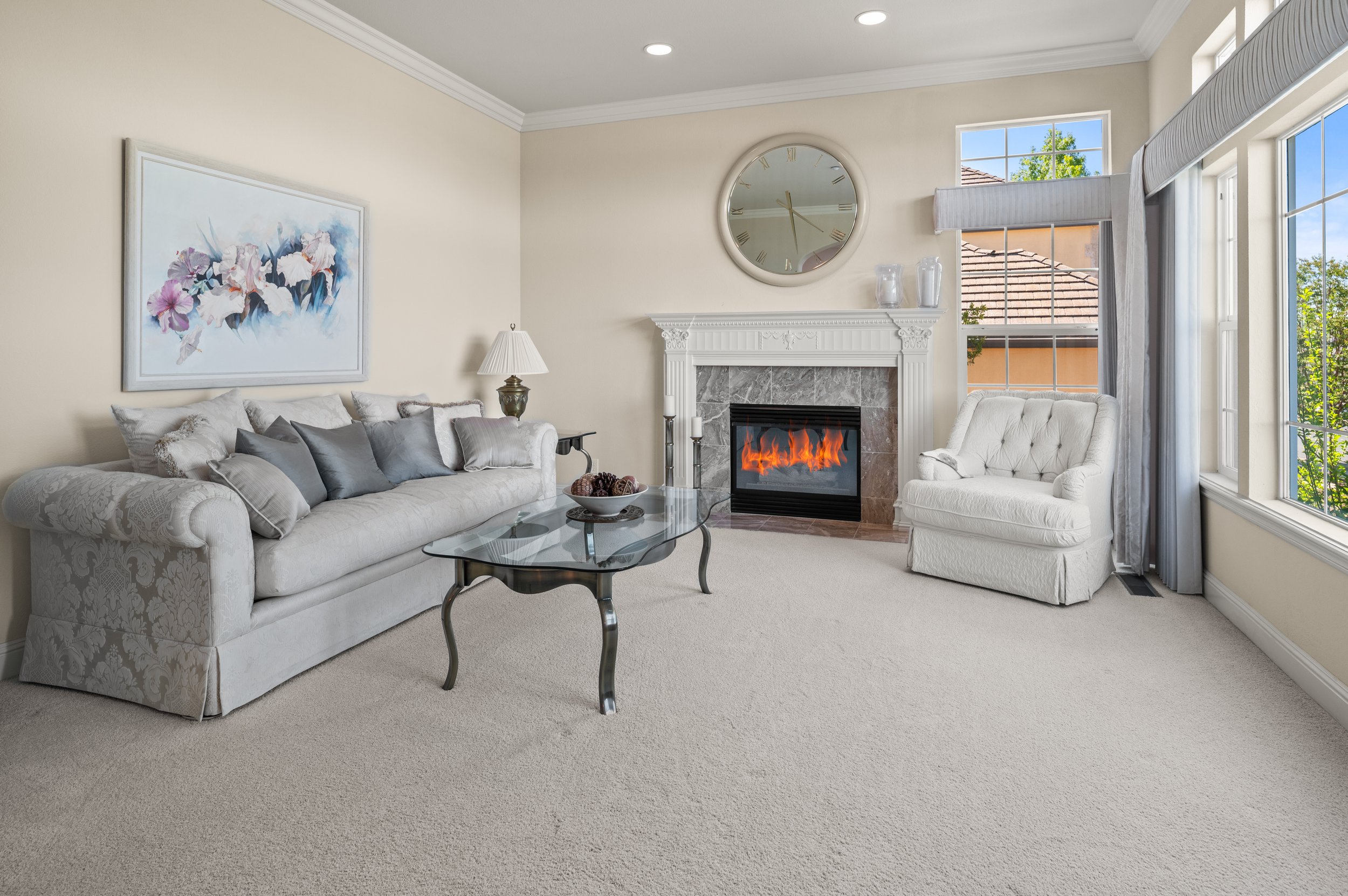 A cozy living room with a white floral-patterned sofa, grey throw pillows, and a glass coffee table with decorative items. There is a white armchair near large windows with blue sky and green trees outside. A fireplace with a marble surround is topped by a round mirror and two vases. A painting of pink and white flowers hangs on the wall, and a table lamp with a beige shade is beside the sofa.