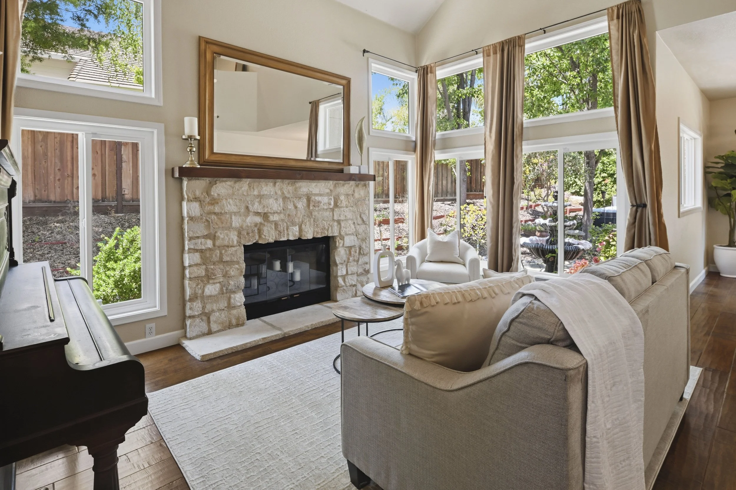 Living room with large windows, a brick fireplace, beige sofa, a white armchair, a wooden coffee table, and a piano, with a view of a backyard garden filled with trees and plants.