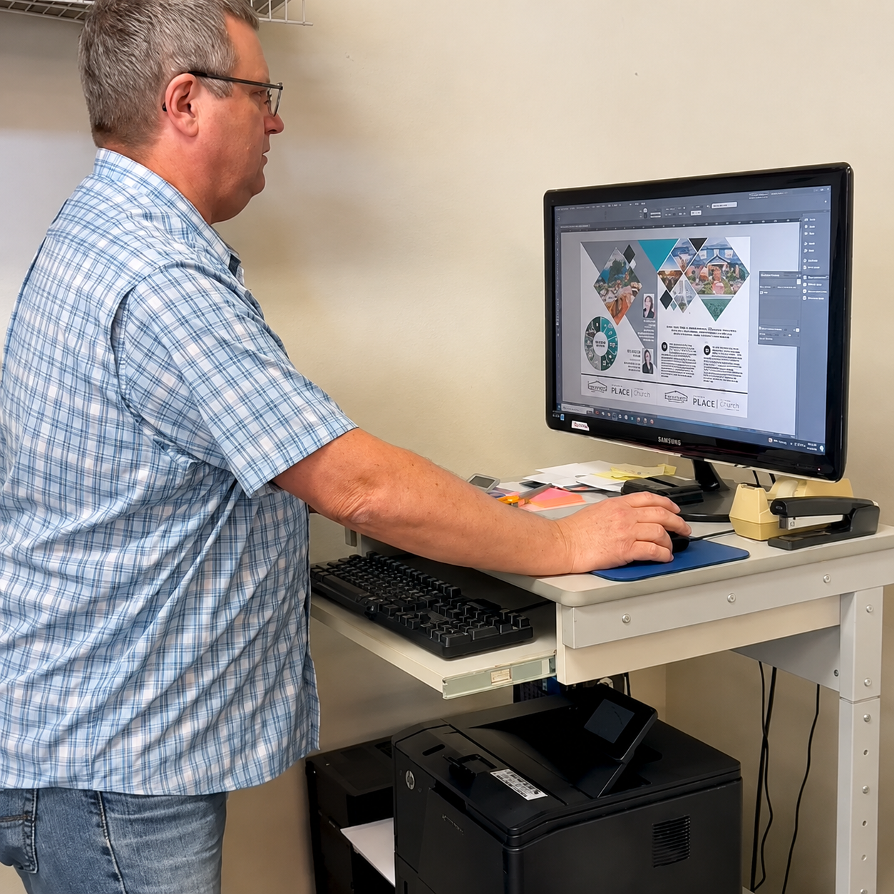Man in plaid shirt working on a computer at a workstation with a monitor, keyboard, and office supplies.
