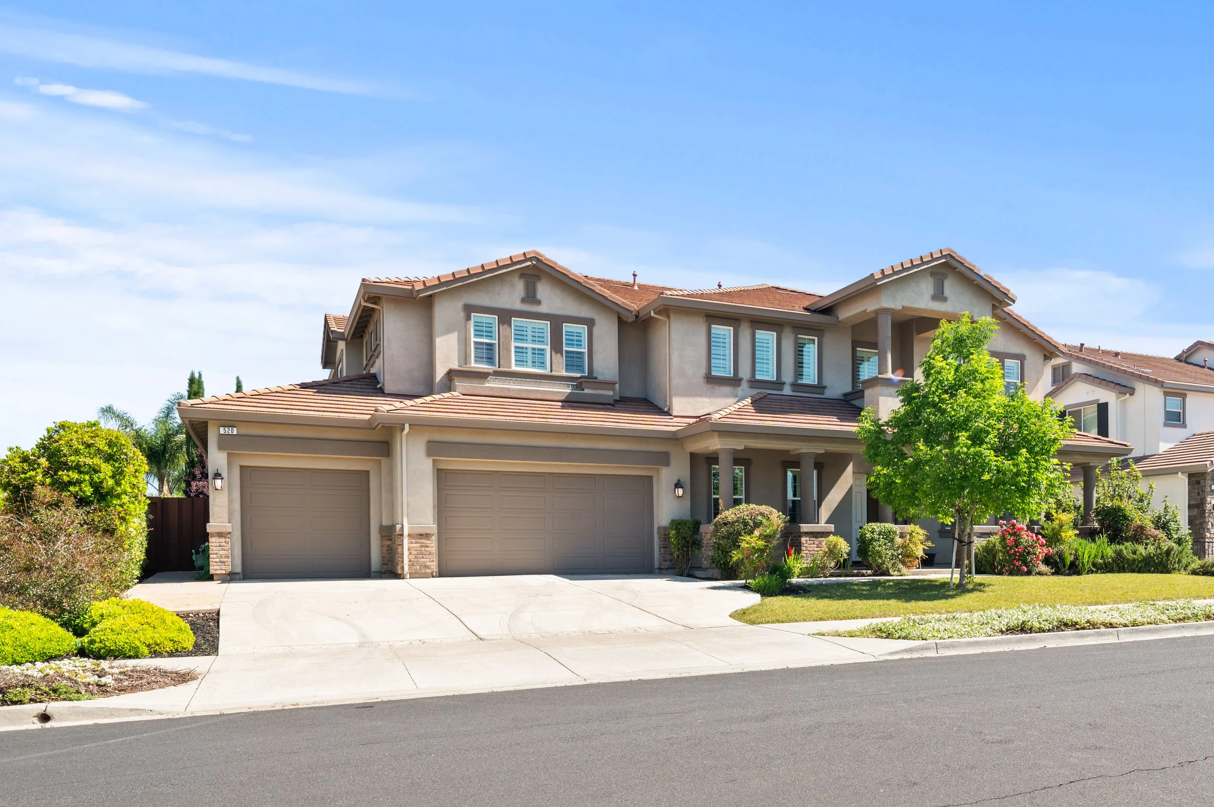 A two-story suburban house with a garage, red tile roof, beige exterior, and well-maintained front yard with plants and small trees, under a clear blue sky.
