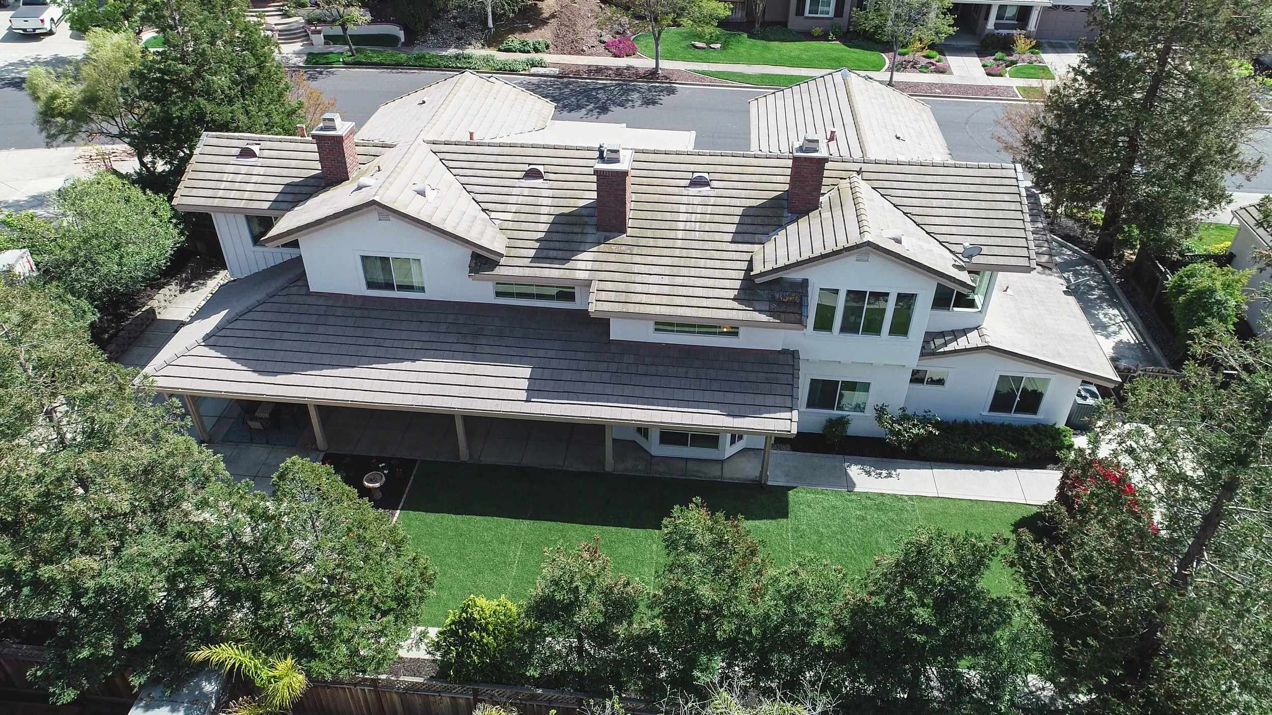 Aerial view of a large white house with a gray tiled roof, multiple chimneys, and large windows, surrounded by green trees and a well-maintained lawn.