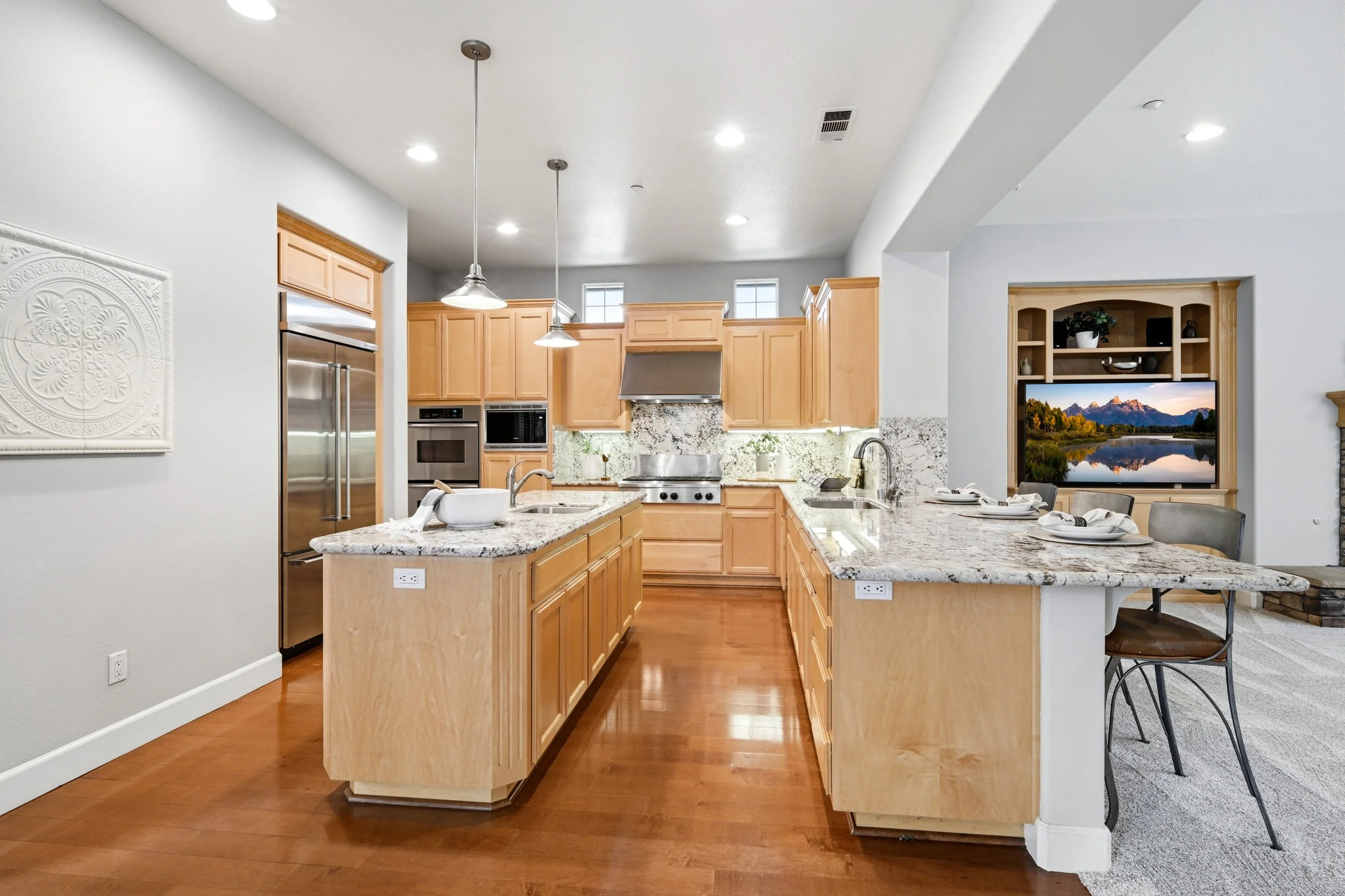 A modern kitchen with light wood cabinets, granite countertops, stainless steel appliances, and a kitchen island with seating. There are recessed ceiling lights, pendant lights over the island, a window above the sink, and a flat-screen TV displaying a landscape scene in the adjacent room.
