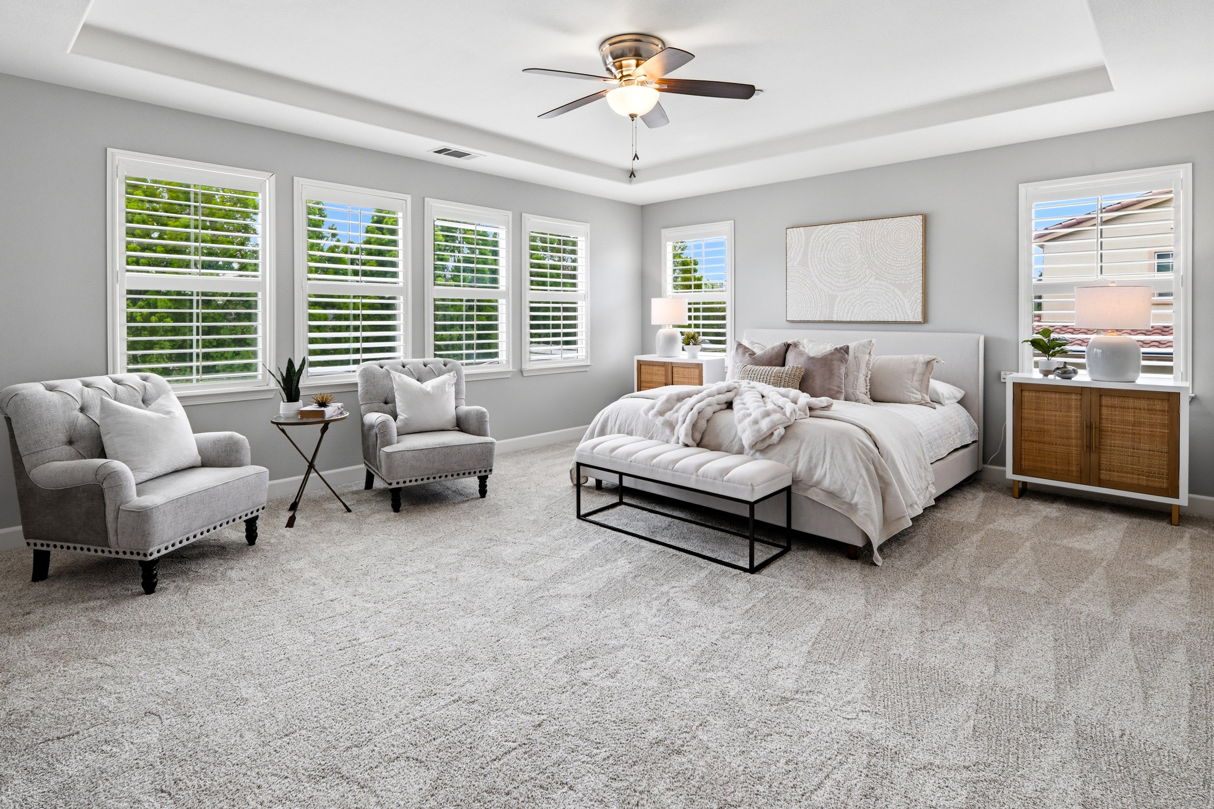 Bright bedroom with multiple windows, a bed with white bedding and decorative pillows, two side tables with lamps, a ceiling fan, and beige carpet.